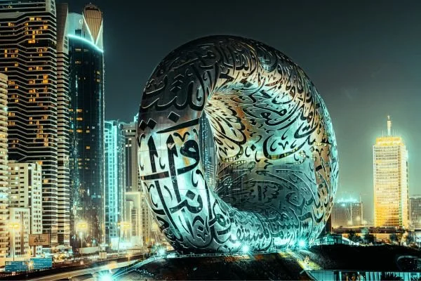 A large, illuminated, donut-shaped building with intricate Arabic calligraphy on its surface, set against a city skyline at night with modern high-rise buildings.