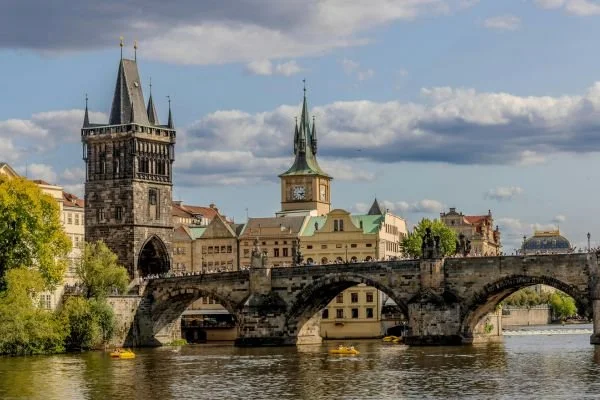 View of Charles Bridge over the Vltava River in Prague, Czech Republic, featuring historic towers and buildings against a partly cloudy sky.