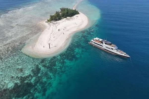 A small tropical island with a few trees, surrounded by clear blue water with coral reefs nearby, and a large yacht anchored close to the shoreline.