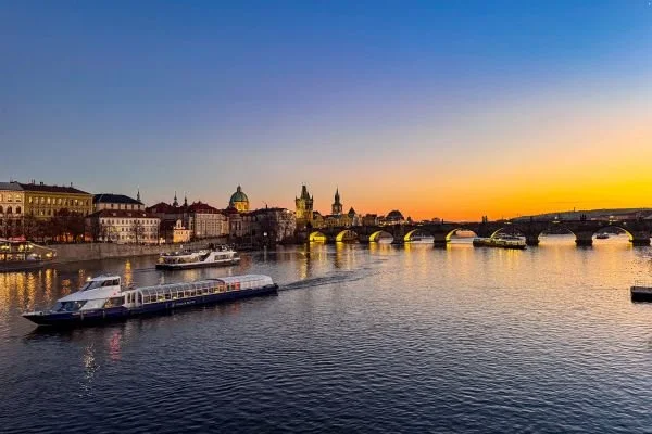 View of a river at sunset with boats on the water and a city skyline with historic buildings and a bridge in the background.