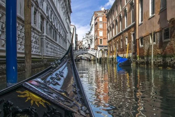 View from a gondola navigating a narrow canal in Venice, Italy, with historic buildings on each side and a small bridge in the distance.