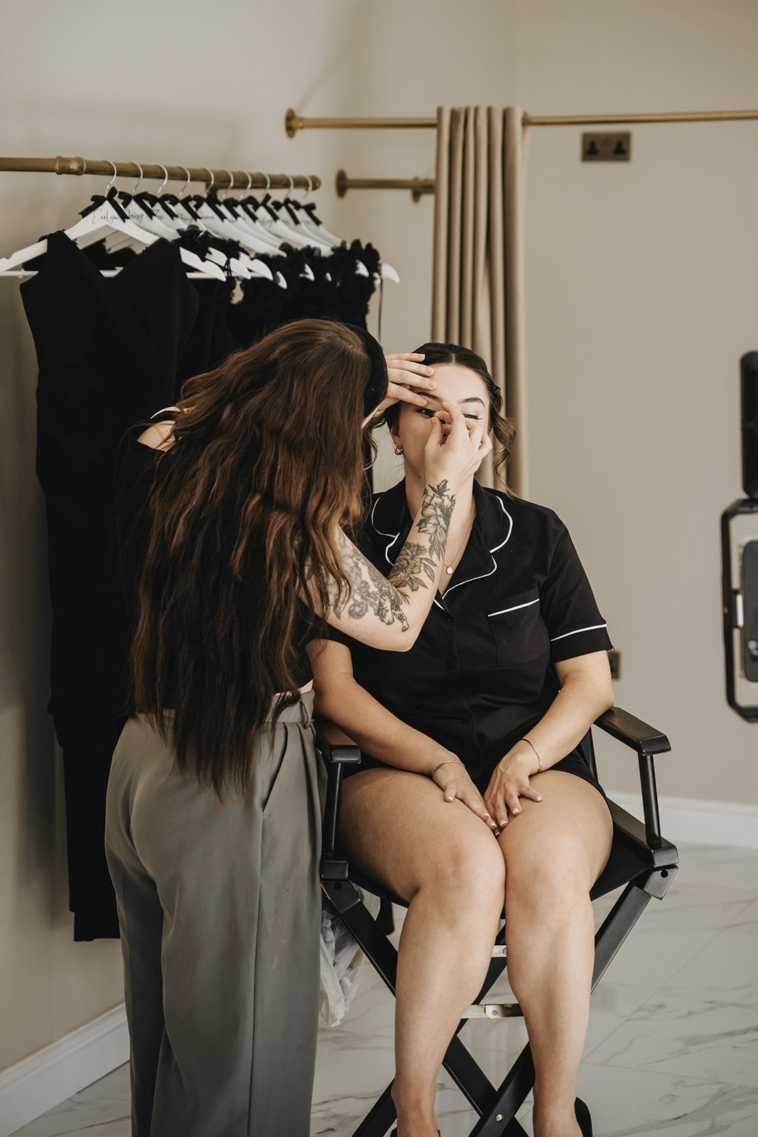 Makeup artist applying makeup to a woman seated in a chair in a room with black dresses hanging on a rack in the background.