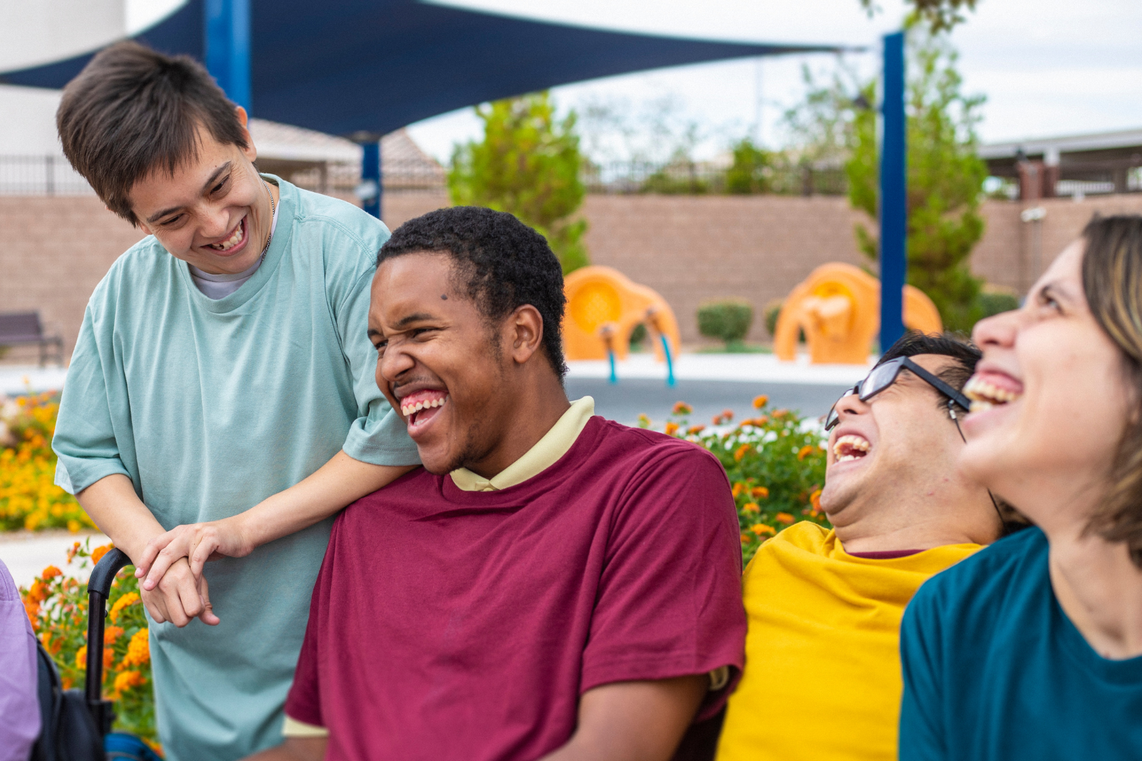 Groupe de cinq personnes souriantes et riant ensemble dans un jardin public, avec des fleurs orange et un remorqueur de jouets en plastique en arrière-plan.