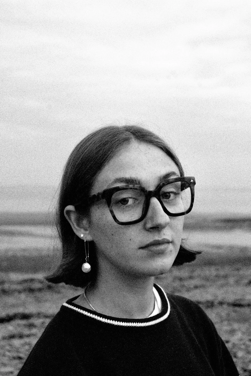A black and white headshot of Emma Morgan. She has short brown hair, freckles, large glasses, dangly earrings and a woolly jumper. She is standing outside on a beach on a cloudy day.