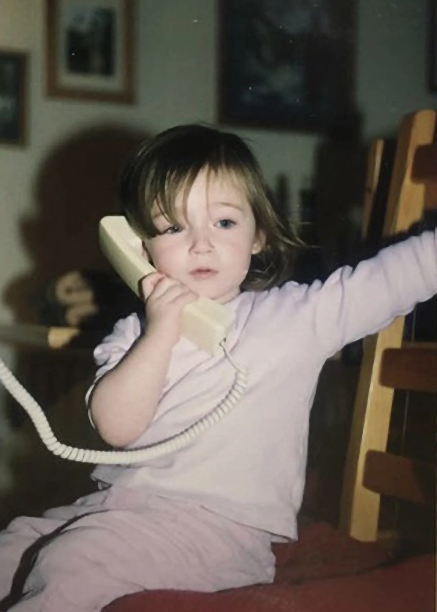A photo of Emma when she was little. She is sitting on a wooden chair and holding a beige landline telephone to her ear.