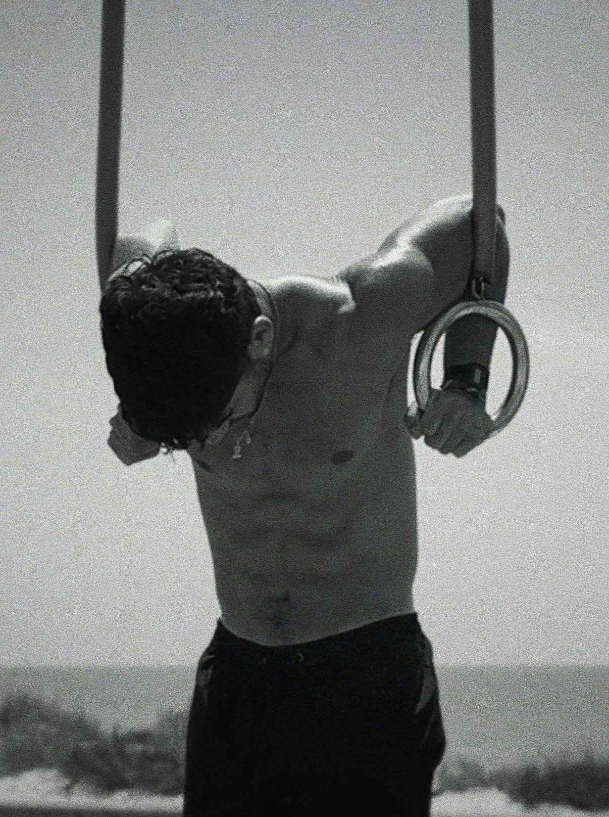 A shirtless man with glasses and headphones, holding gymnastic rings, doing an exercise outdoors with a scenic view in the background.