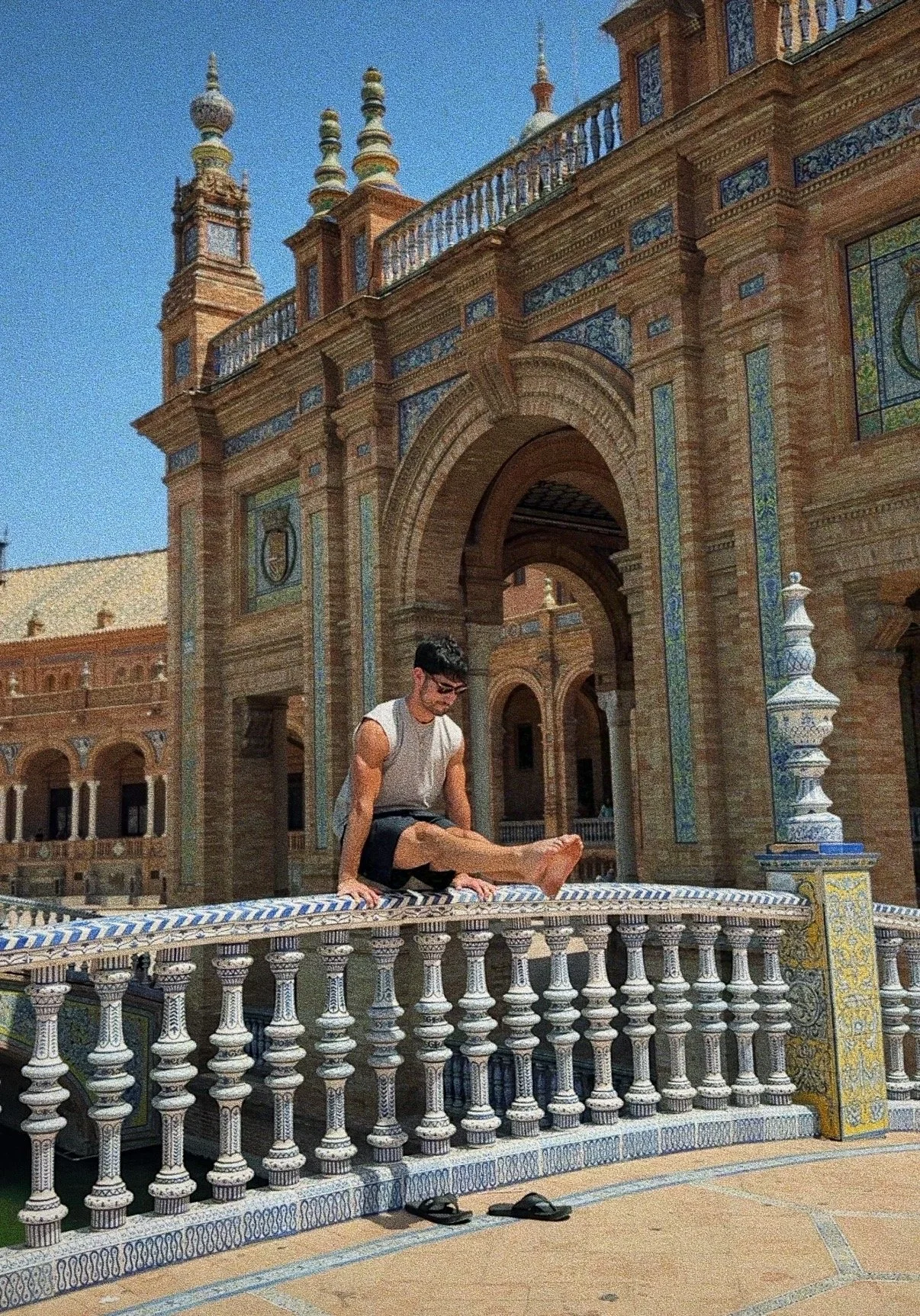 A young man wearing glasses, sleeveless shirt, and shorts, sitting on a decorative tiled balustrade in front of a historic building with arches, towers, and ornate blue and yellow tilework on a sunny day.