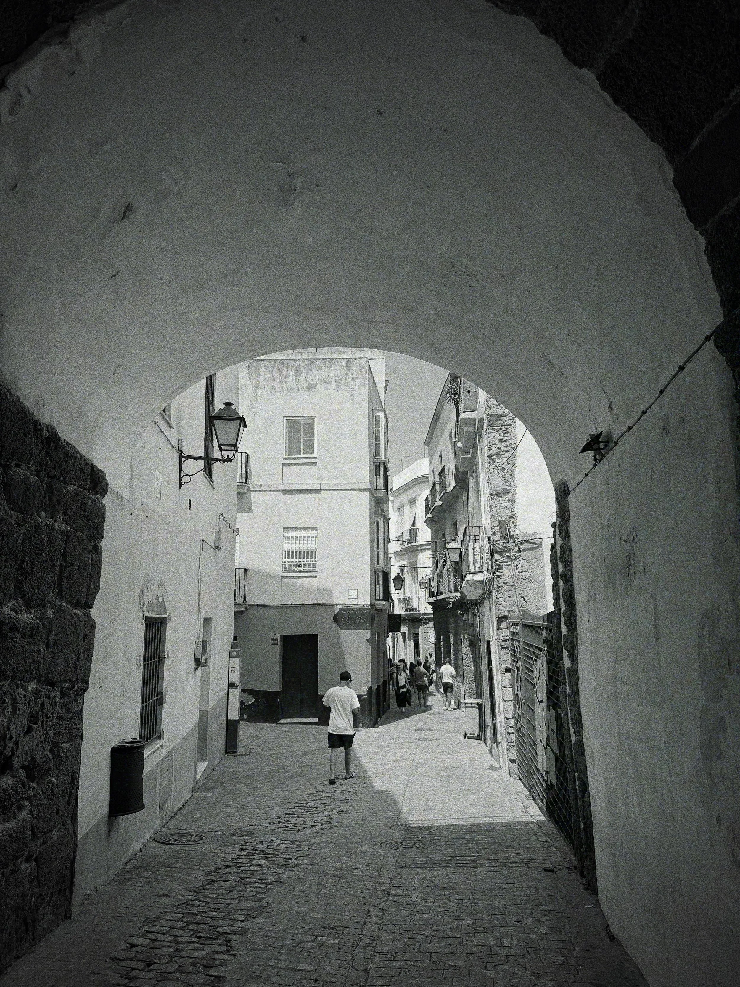 Black and white photograph of a narrow, cobblestone street viewed through an archway in an old European town, with people walking along the street, buildings with balconies, and street lamps.