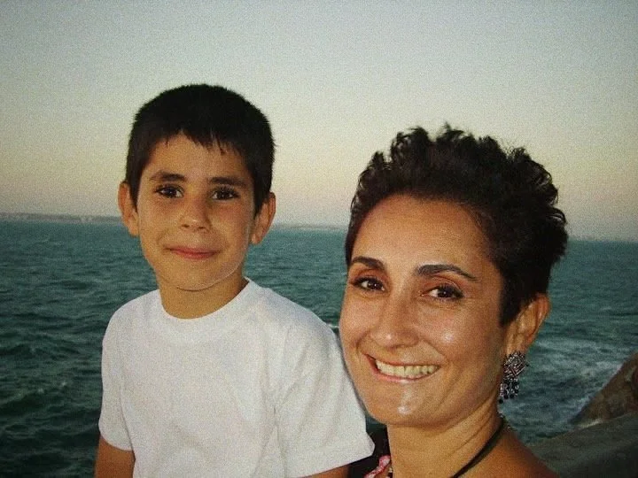 A woman and a boy smiling together near a body of water, possibly the ocean, during sunset or sunrise.