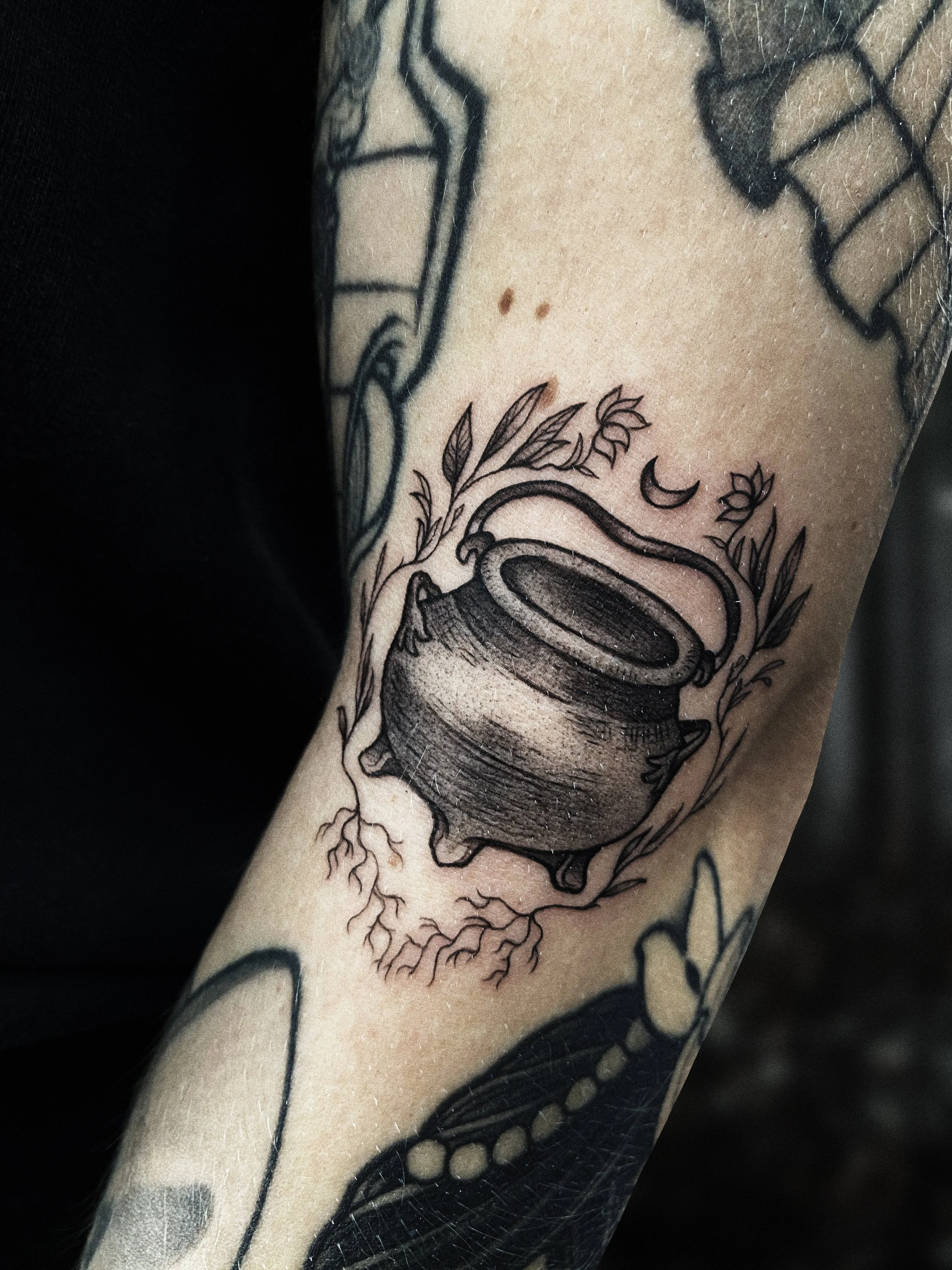 Tattoo of a cauldron with a small crescent moon above it, surrounded by leaves and roots.