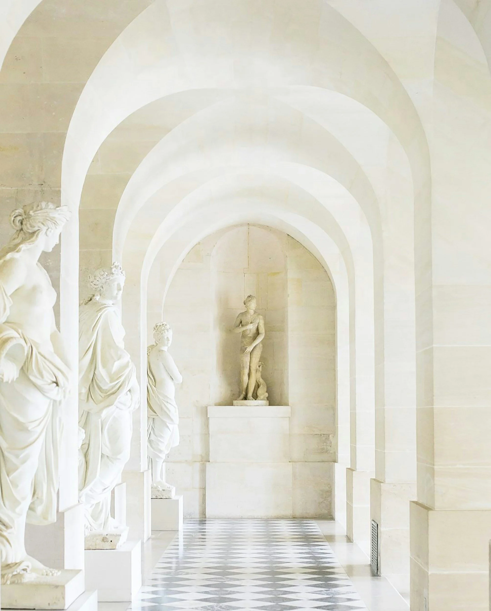 A hallway with a checkered black-and-white floor, vaulted ceilings, and white marble statues of women along the left side, with a central statue of a woman at the far end.