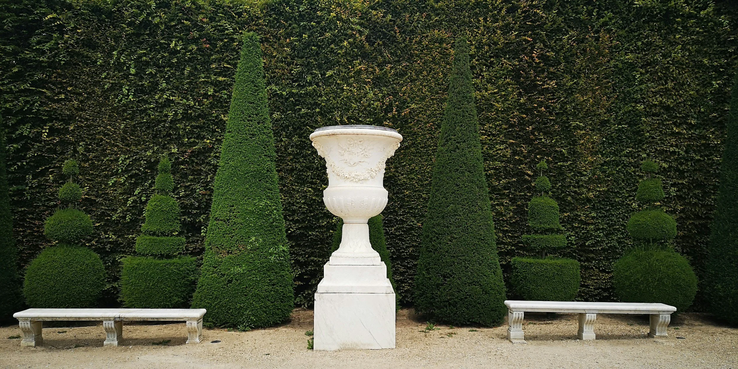 A white marble urn on a pedestal flanked by two white marble benches, with manicured cone-shaped and spherical shrubs in the background.