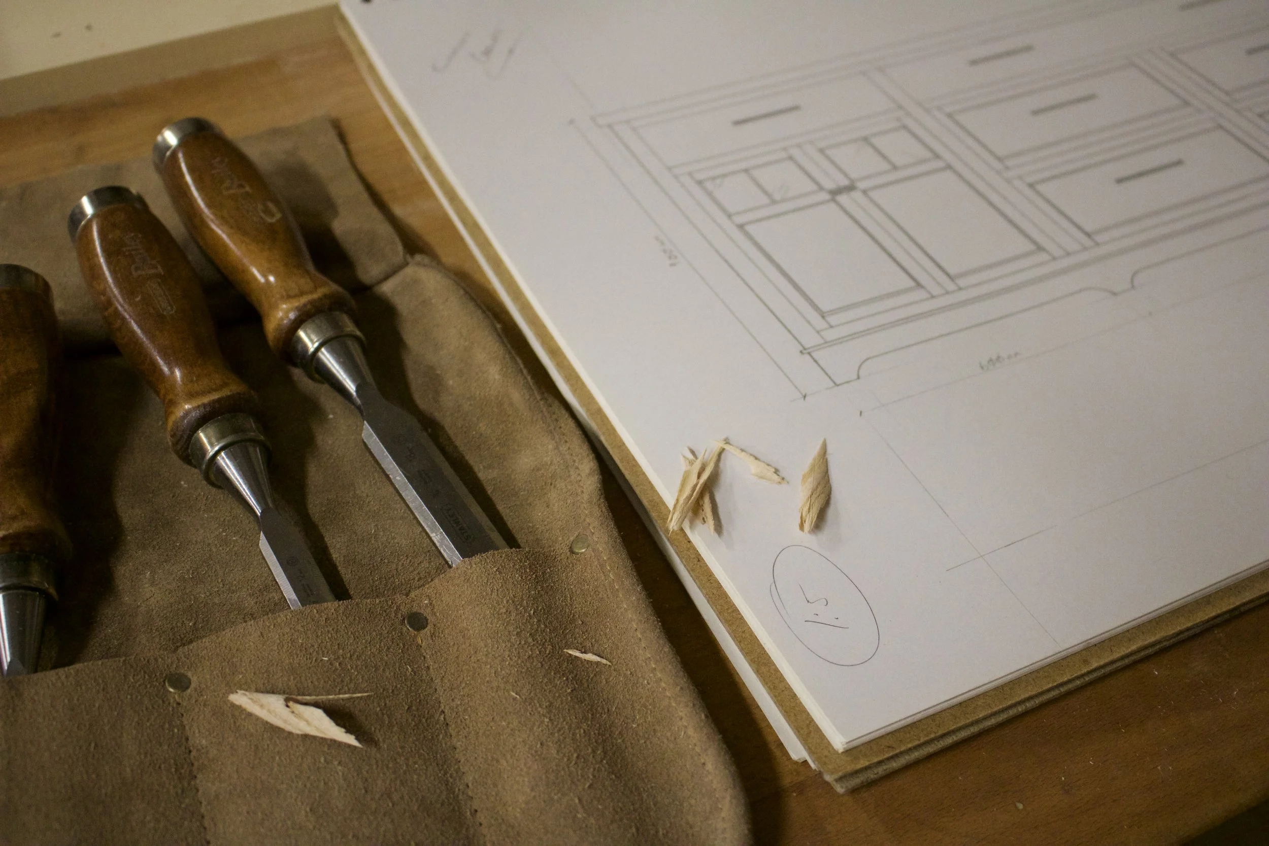 A set of woodworking chisels with wooden handles resting in a brown fabric tool holder on a wooden surface. Next to them is a large paper with a detailed architectural drawing of a window or cabinet and some small wood shavings on top.