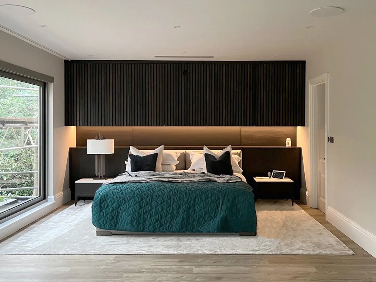 Modern bedroom with fitted furniture and a king-size bed, nightstands with a lamp and a tablet, large sliding glass door with view of greenery.