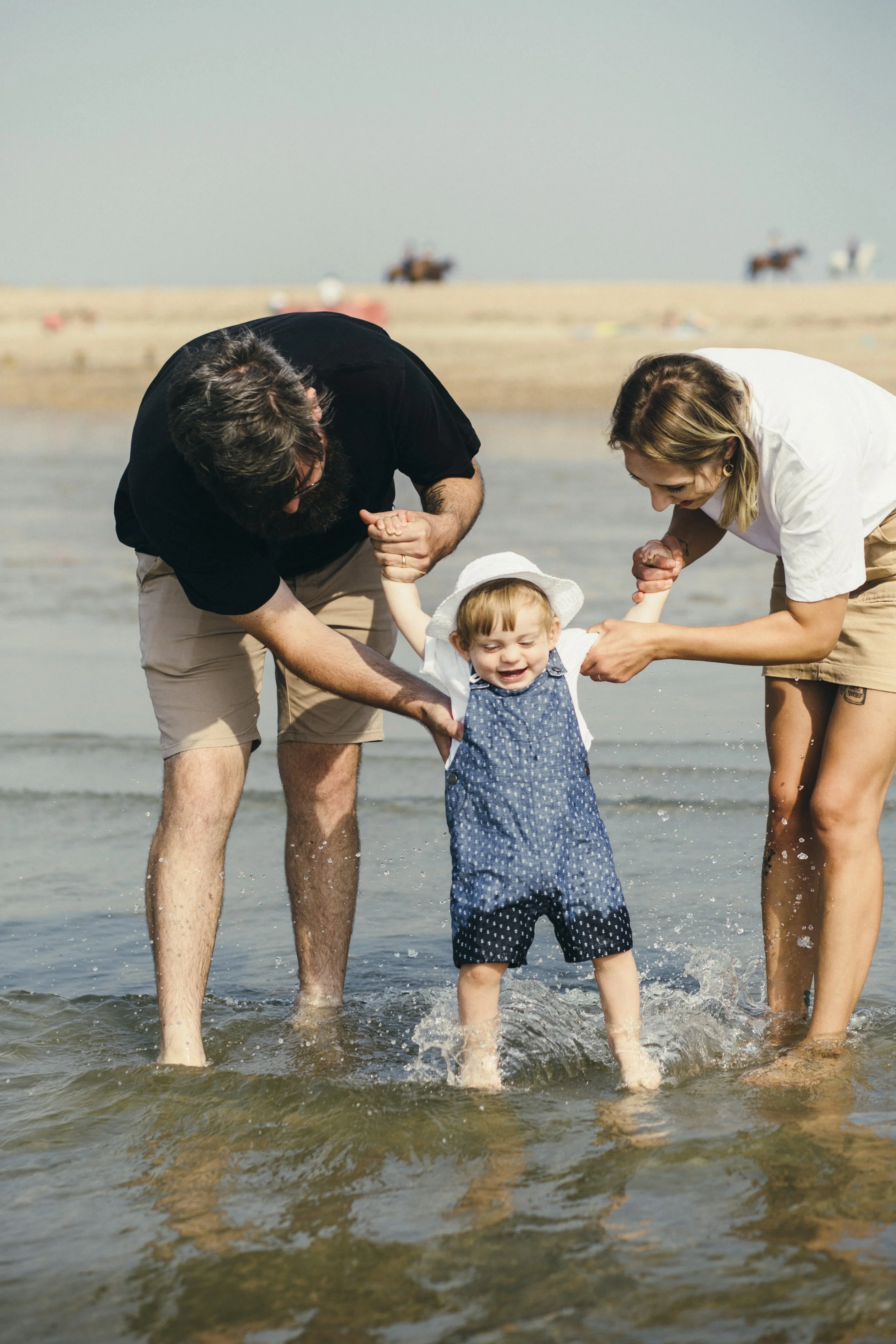 A family of three enjoying time together at the beach, with the parents helping their young child stand in shallow water, smiling and having fun.