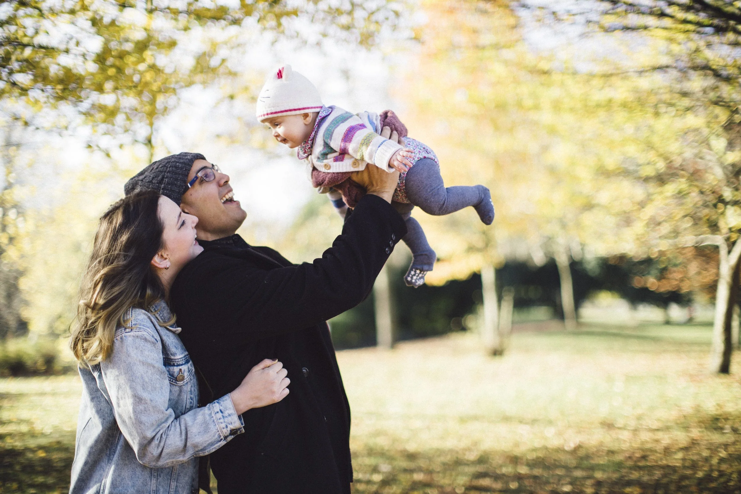 A family enjoying fall in a park: a man lifting a baby girl, supported by a woman.