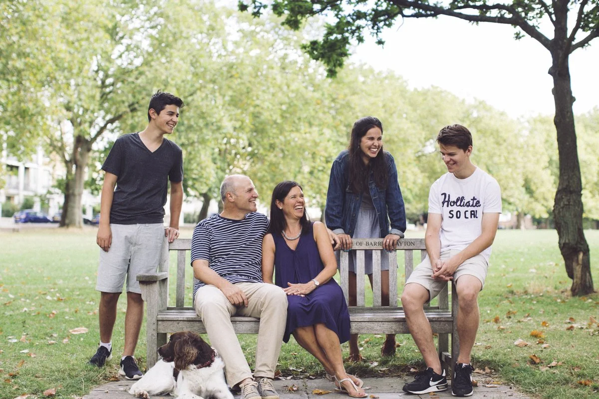 A family of six, including a dog, sitting and standing on a park bench, laughing and enjoying each other on a sunny day with green trees in the background.