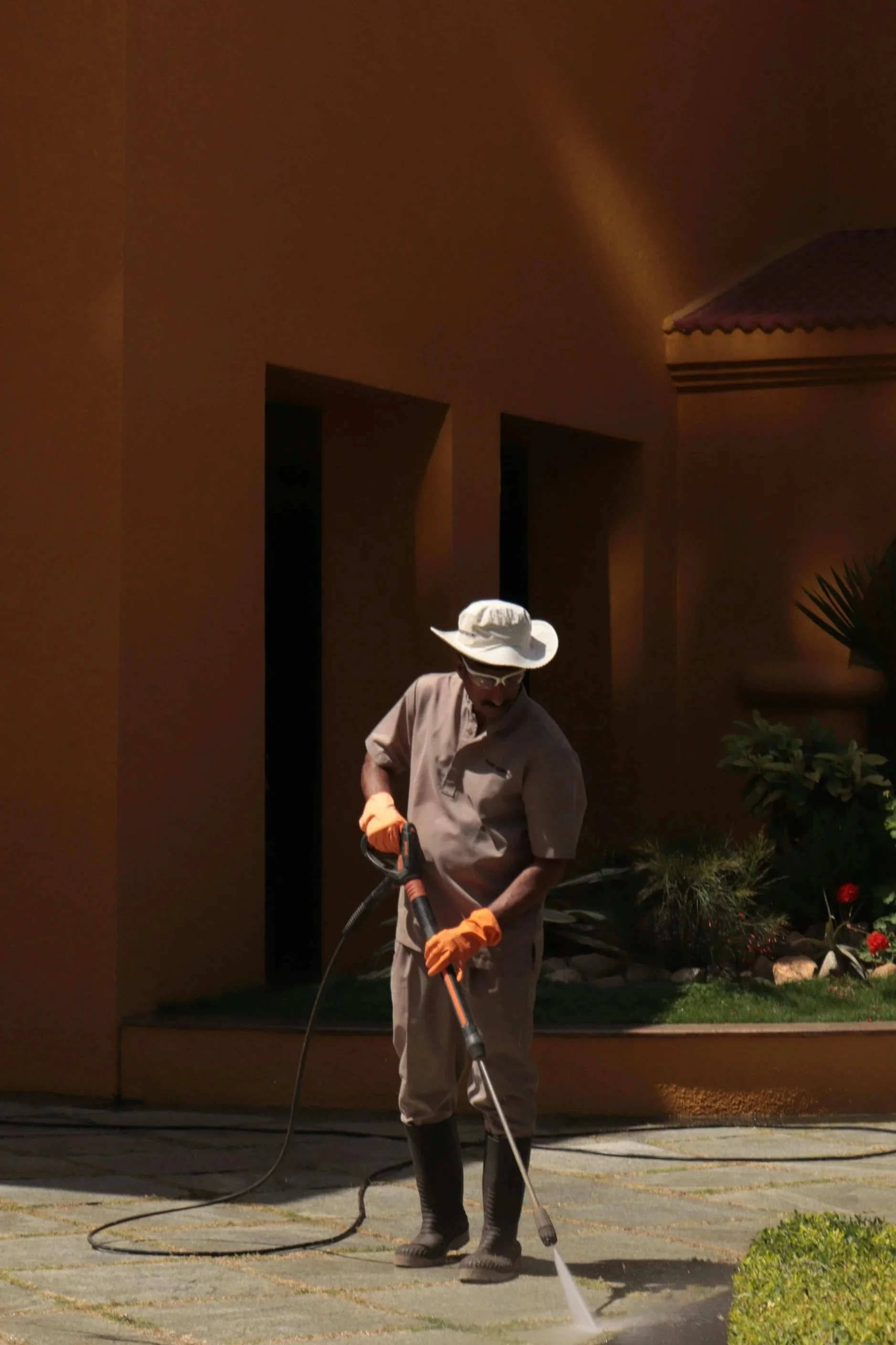Person wearing a wide-brimmed hat, orange gloves, and boots, using a power washer to clean a concrete patio outside a building.