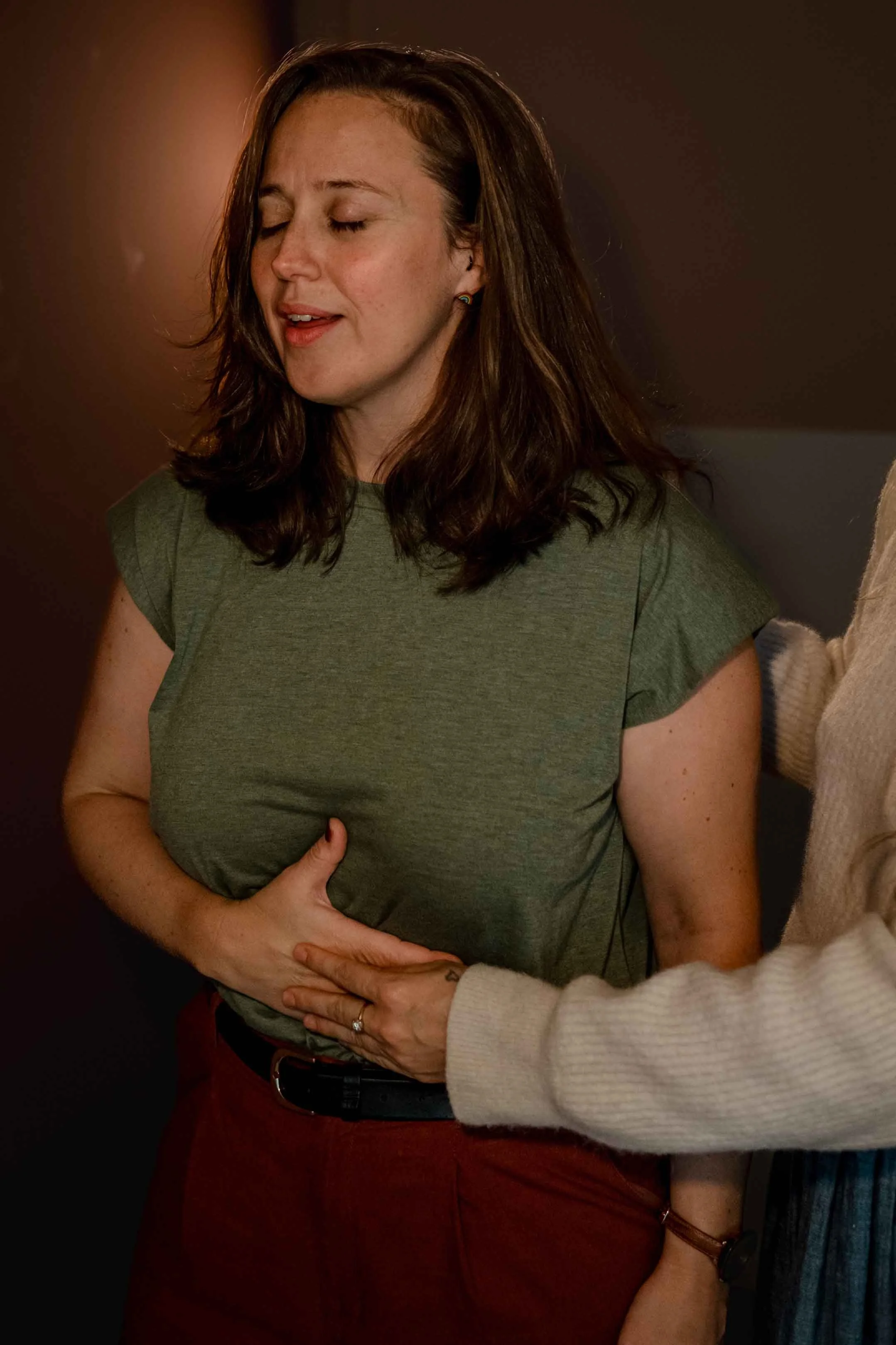 A woman with brown hair, wearing a green t-shirt and maroon pants, practices somatic singing with guidance from Meggy Tucker.