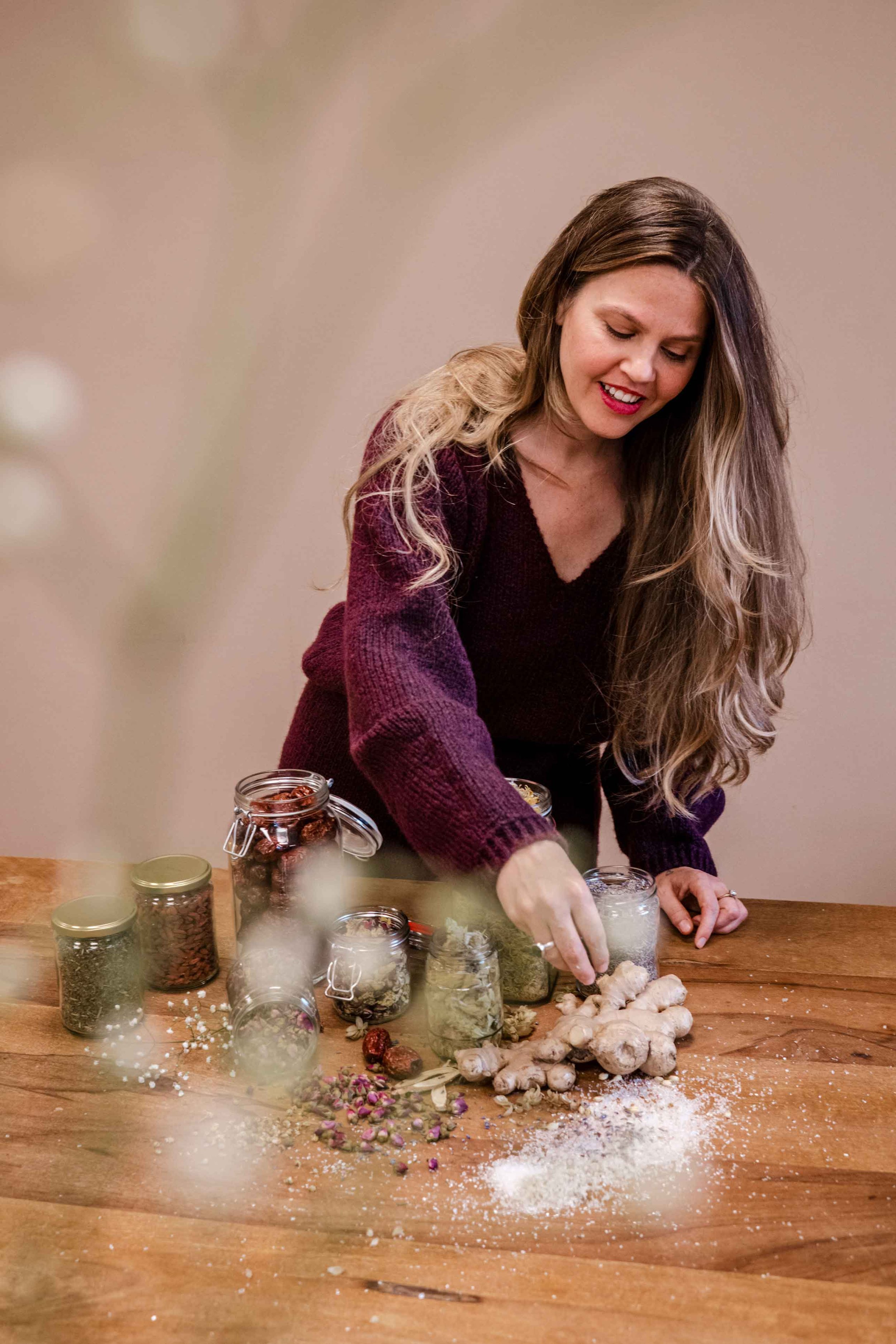 Acupuncturist Meggy Tucker is working with ginger and various dried herbs and spices on a wooden table, surrounded by jars of ingredients.