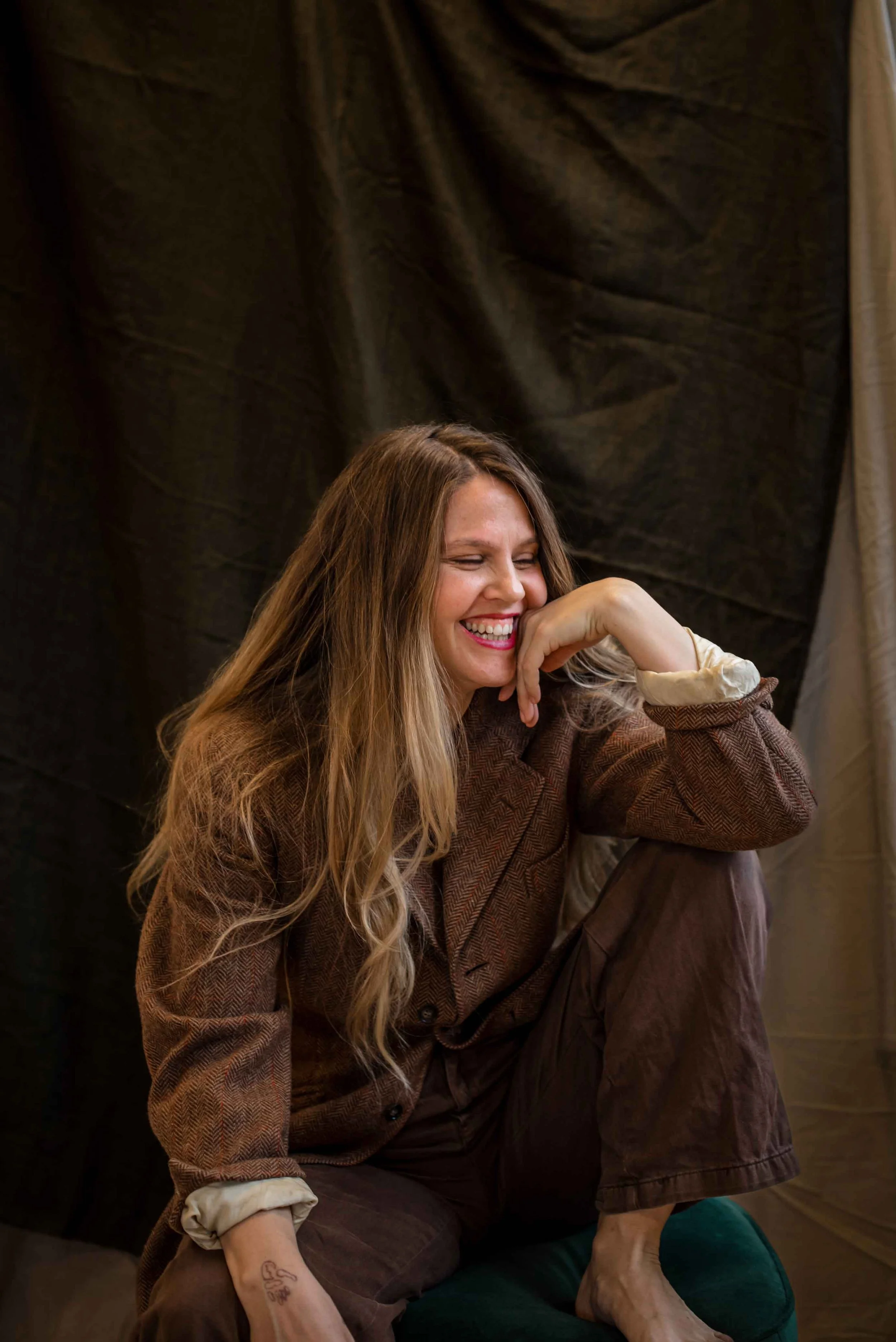 Meggy Tucker is sitting on a dark green stool, smiling with her face leaning on her hand, against a dark background.