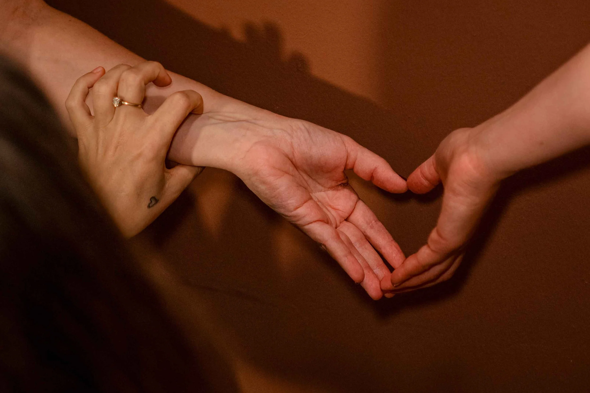 Two hands gently touching, one hand with a wedding ring, set against a brown background.
