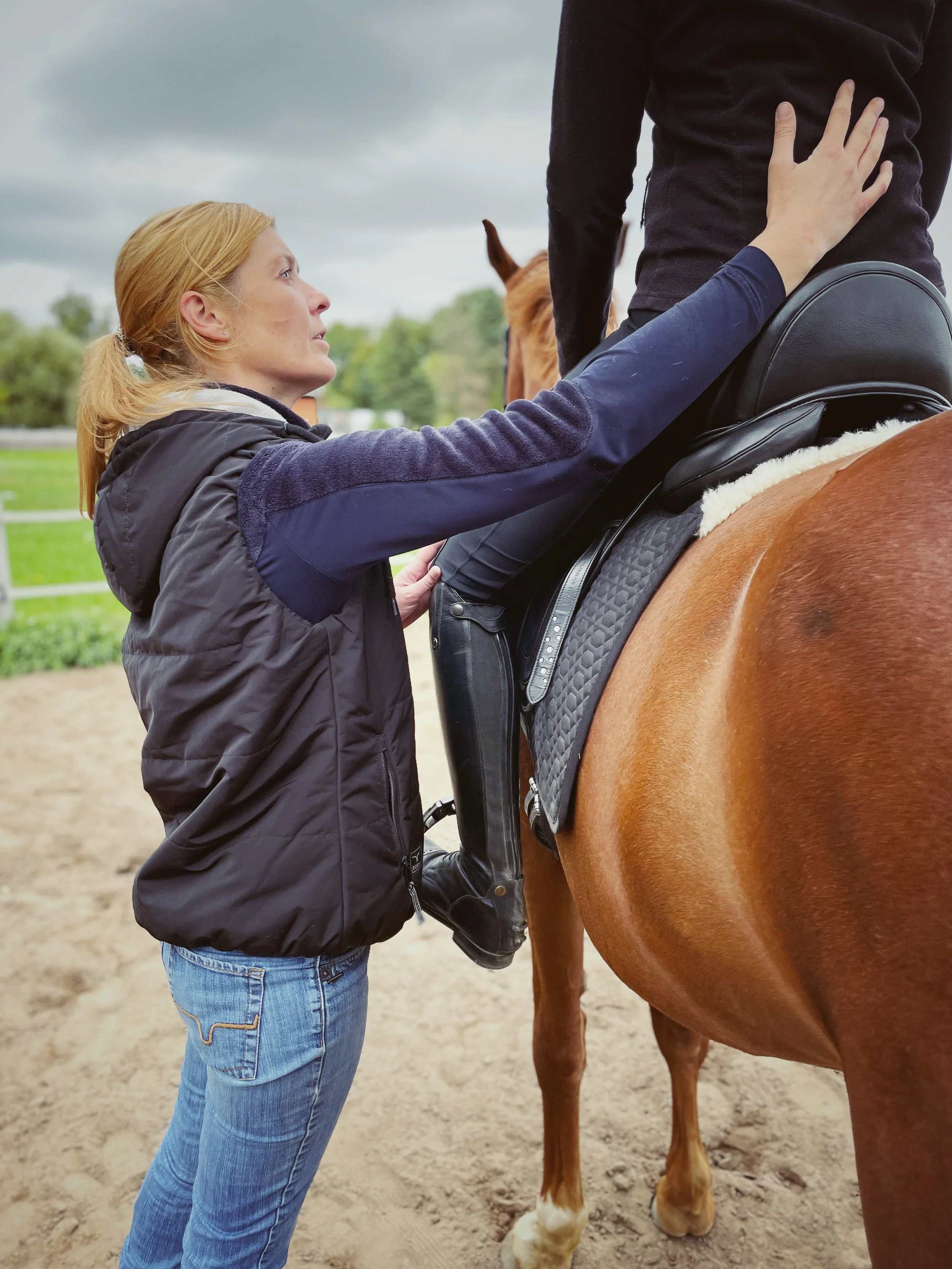 Eine Frau hilft einer Person, die auf einem Pferd reitet, beim Anlegen des Sattels, draußen auf einer Reitanlage.