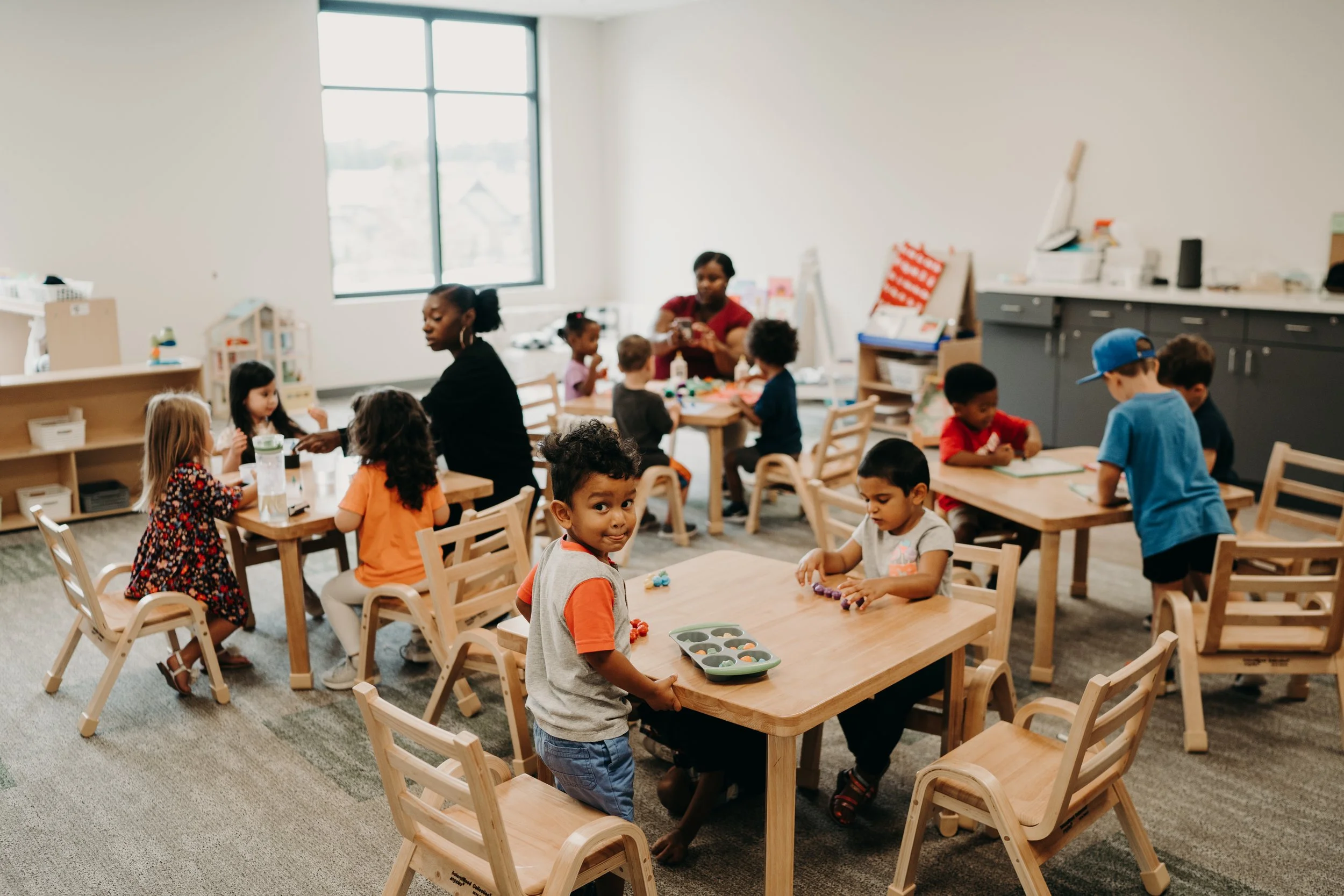 Young children in a classroom playing with toys and engaging in activities at tables, with two adult women supervising.