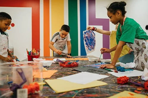 Children and an adult painting and drawing on paper at a colorful art table.