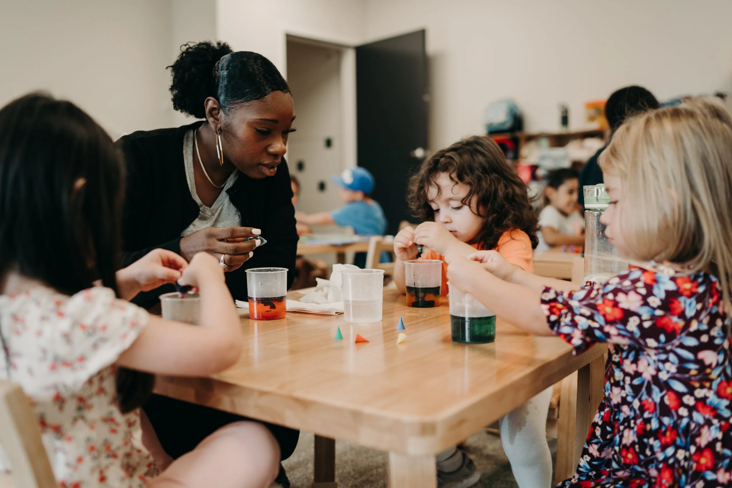 A teacher or caretaker demonstrates a science experiment with children at a wooden table, involving cups with colored liquids and small objects, in a classroom setting.
