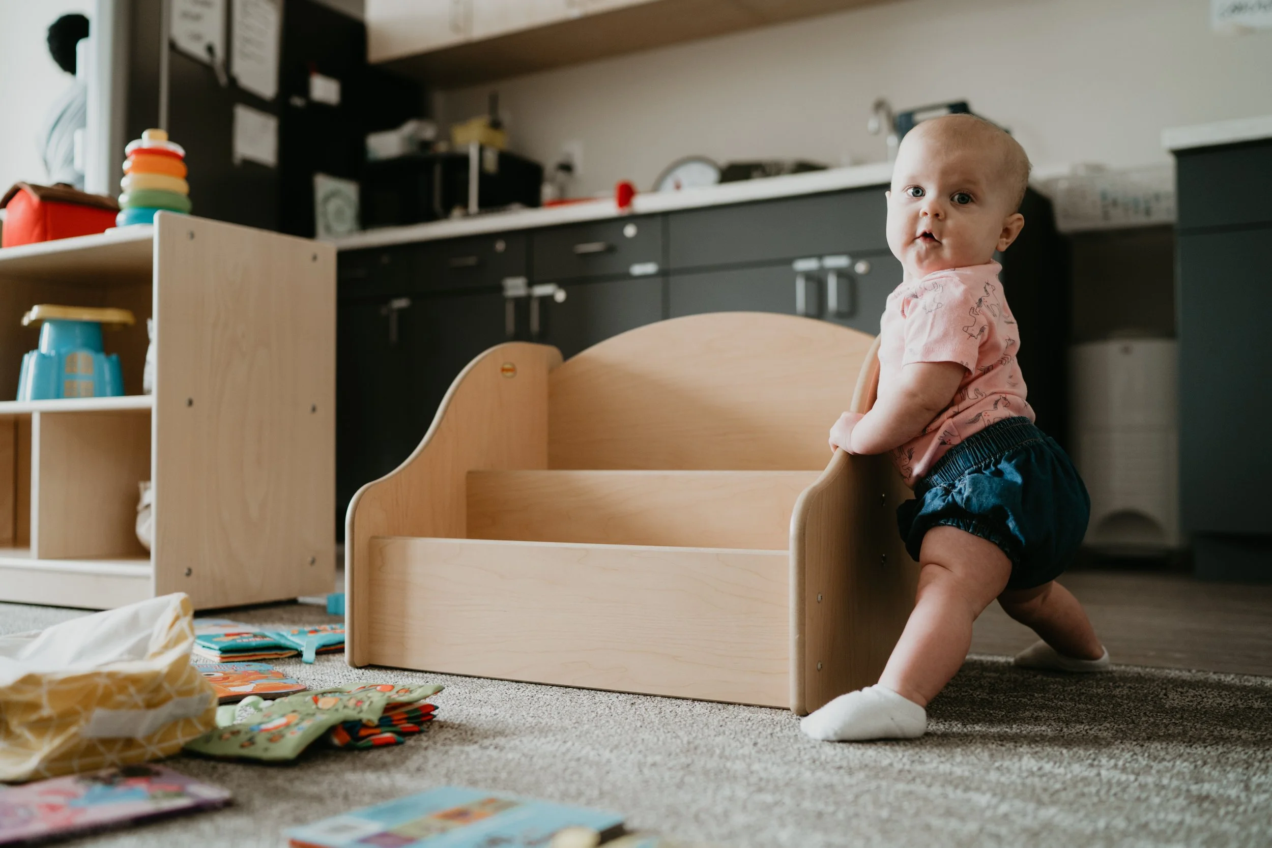 A baby learns to stand with support in a playroom, surrounded by toys and furniture.
