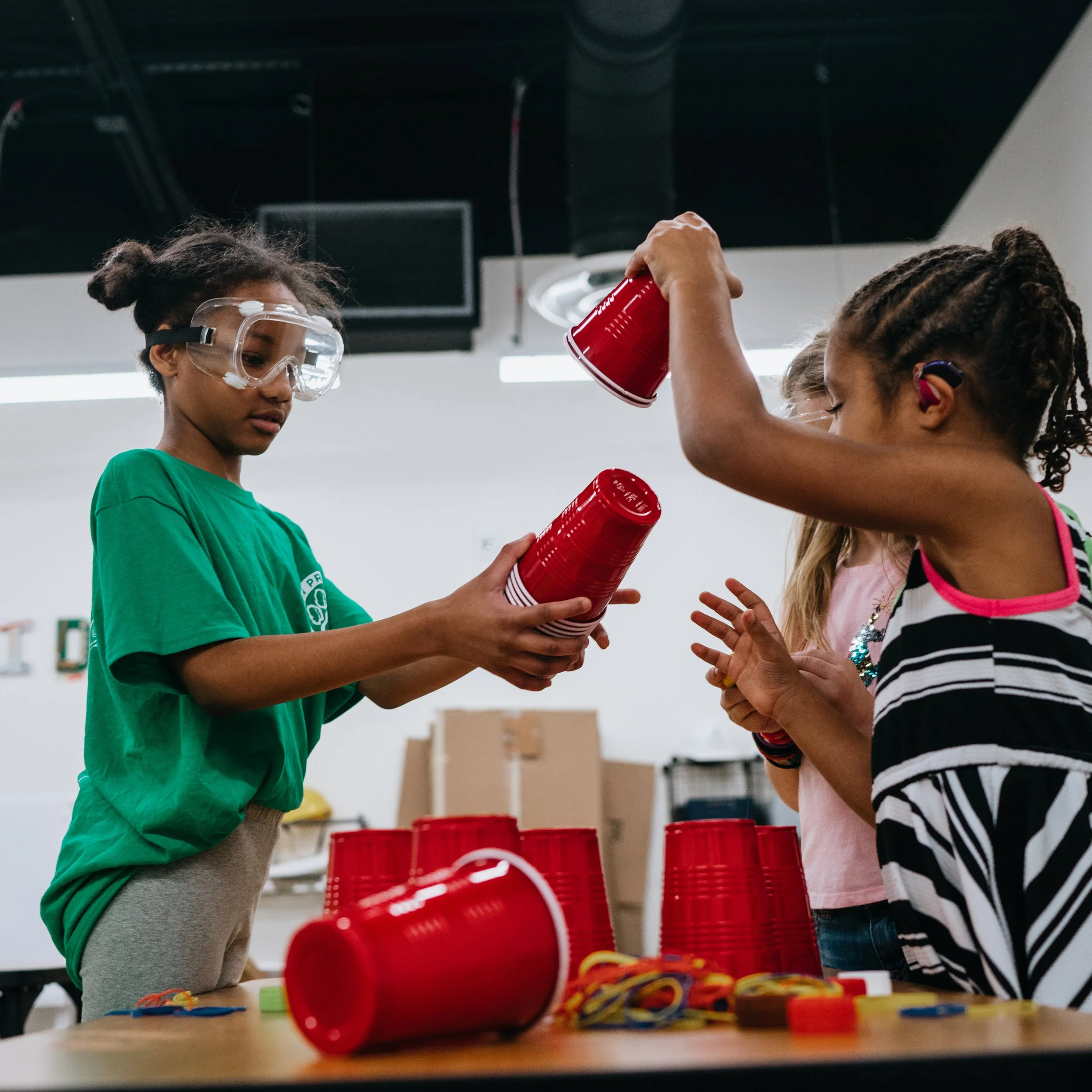 A girl in a green shirt and safety goggles handing red plastic cups to a girl in a black and white striped shirt in a classroom or activity space.