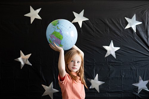 A young girl with red hair holding an inflatable globe above her head, standing in front of a black backdrop decorated with white stars.