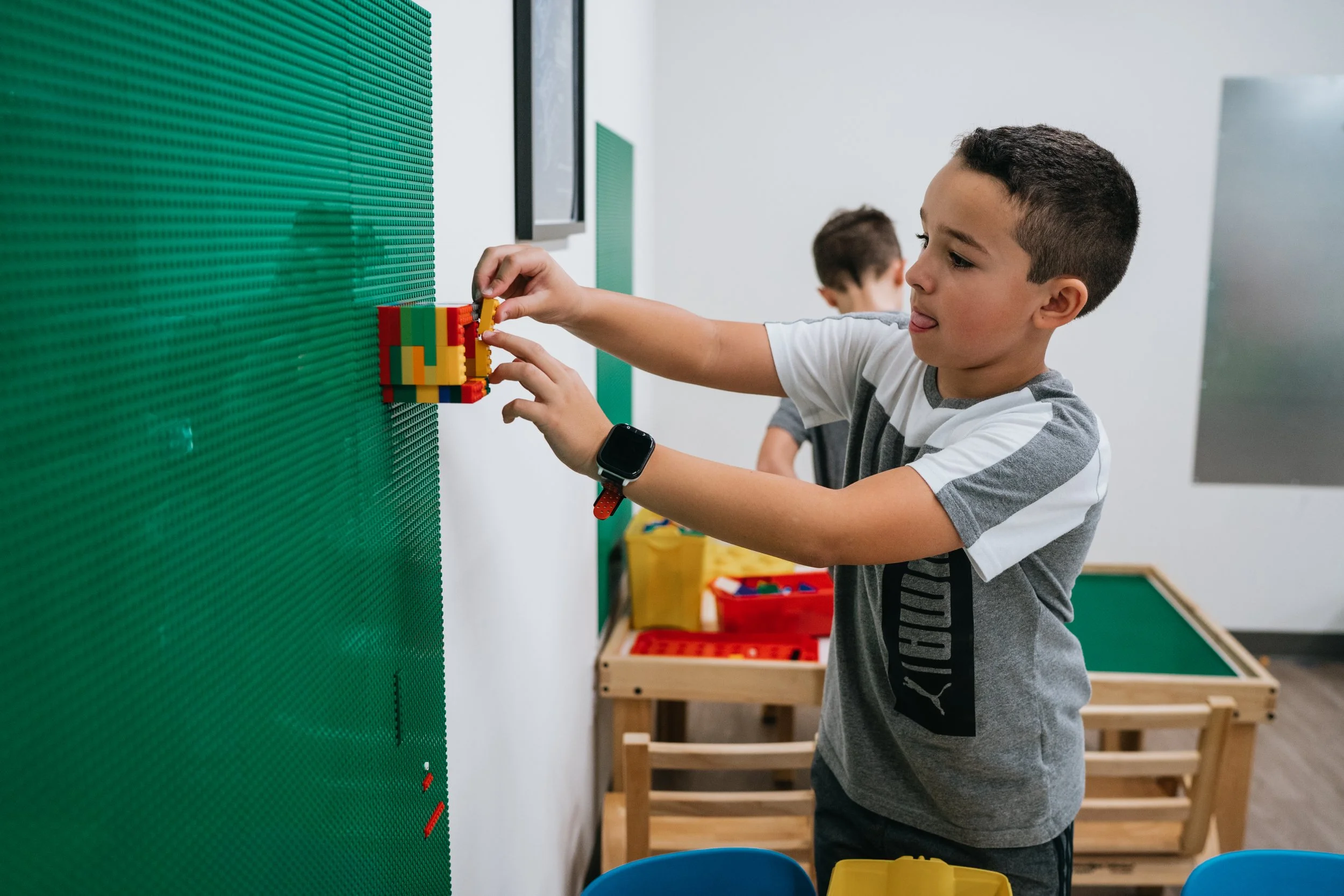 A young boy building with colorful LEGO bricks on a green pegboard wall in a classroom.