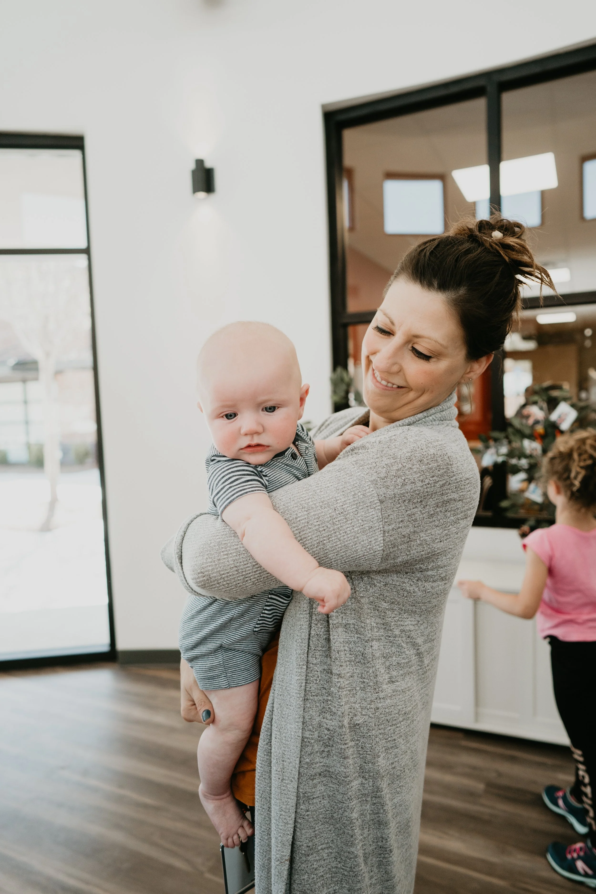 A woman holding a baby inside a modern home with a white interior, large windows, and a wooden floor. A young girl is in the background.