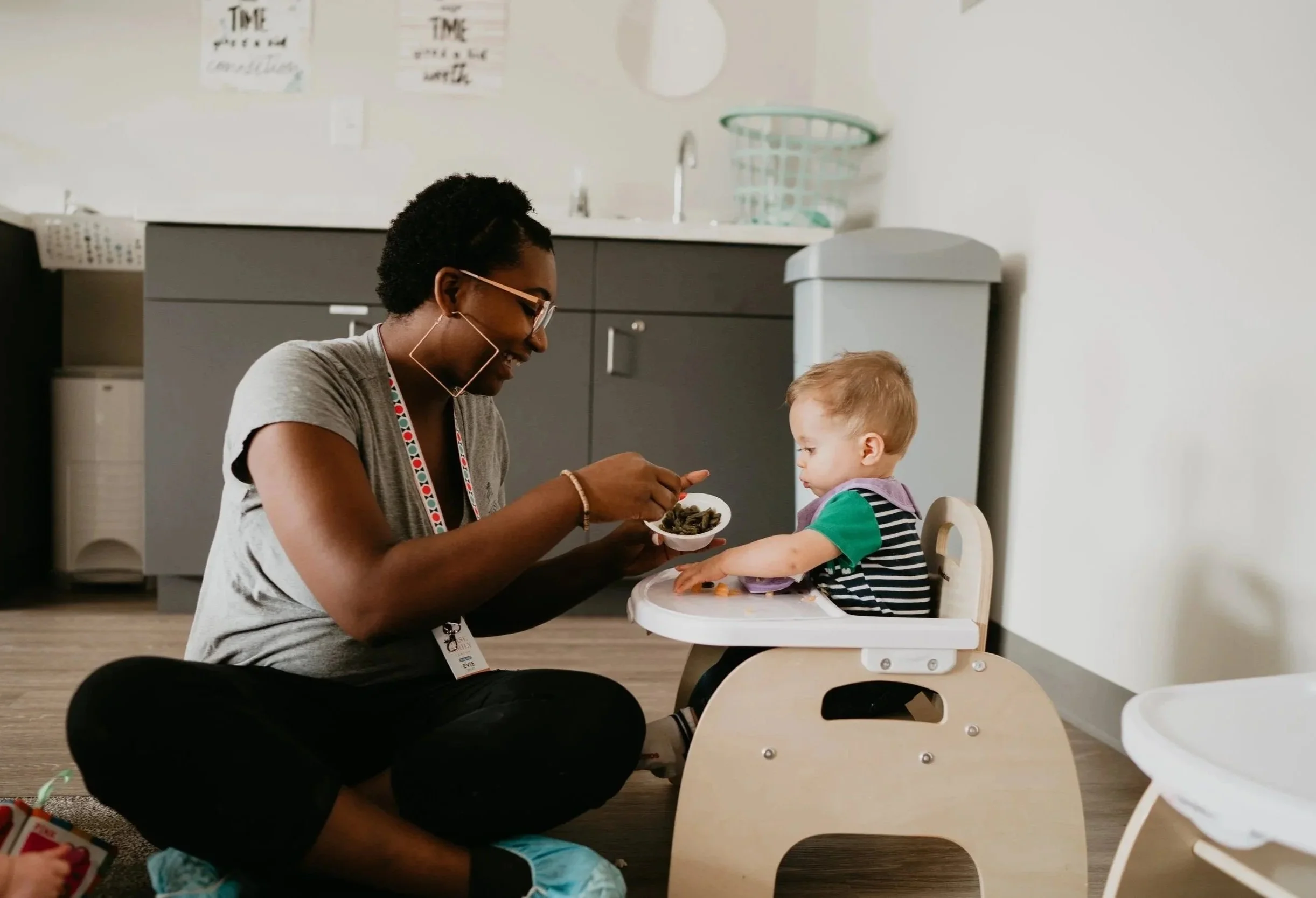 A woman with glasses and earrings feeding a young boy in a high chair in a kitchen or daycare setting.