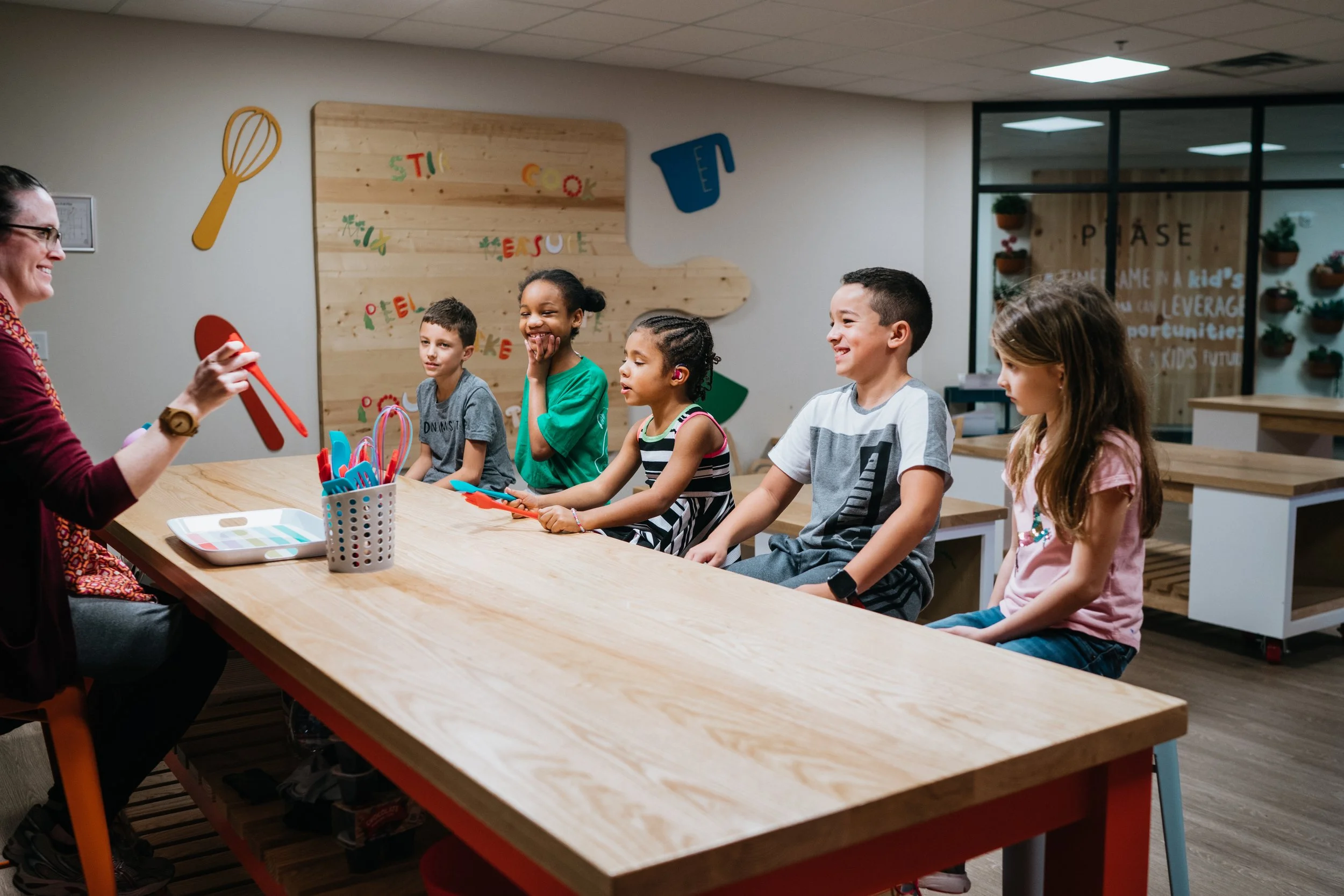 A woman leading a children's cooking class at a wooden table with five children seated, holding colorful cooking utensils, smiling and paying attention.