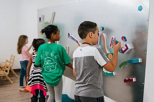 Children drawing on a large gray whiteboard with colorful 3D letters and shapes