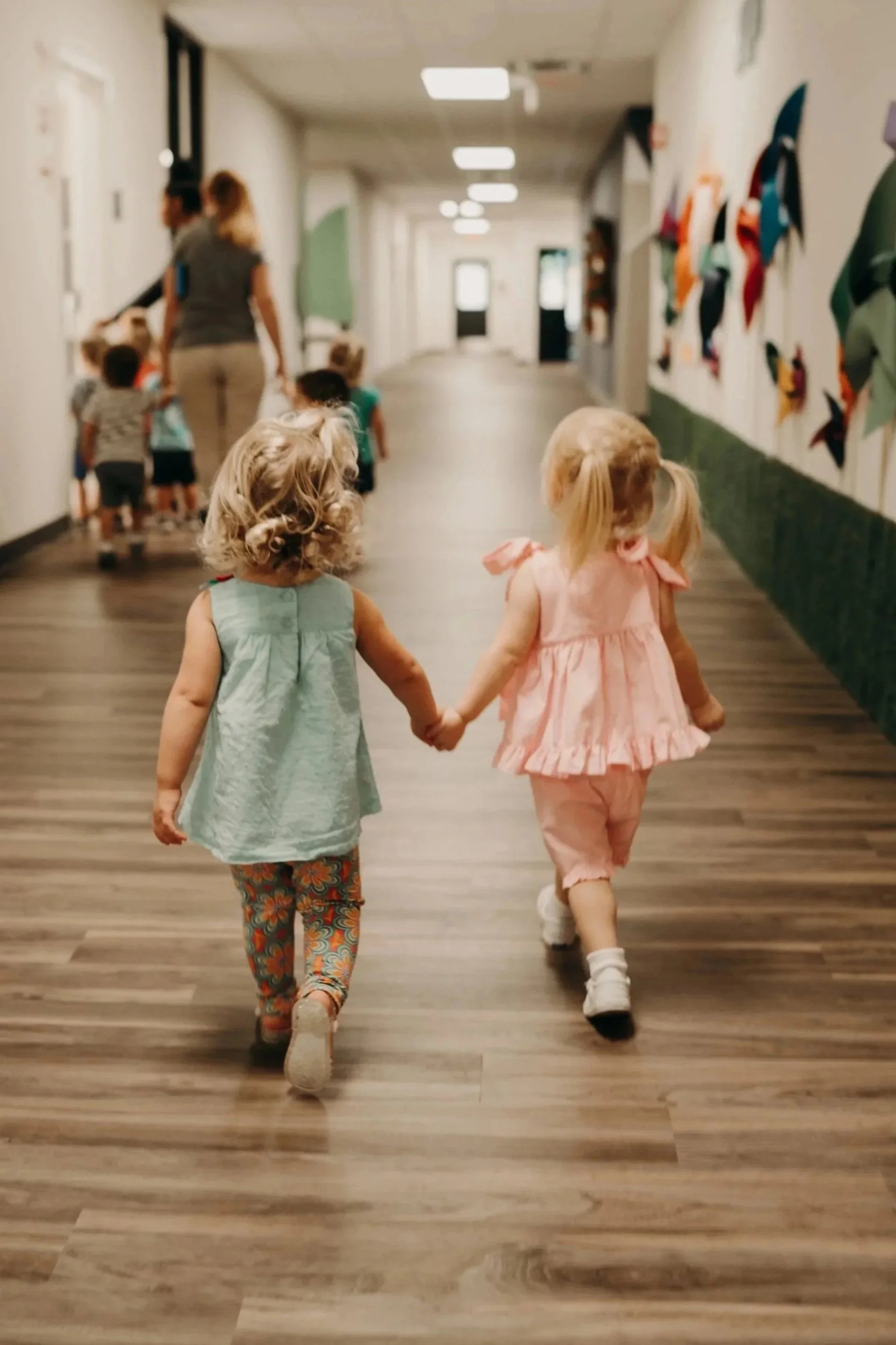Two young girls holding hands and walking down a hallway with colorful artwork on the wall. Other children and a caregiver are visible in the background.