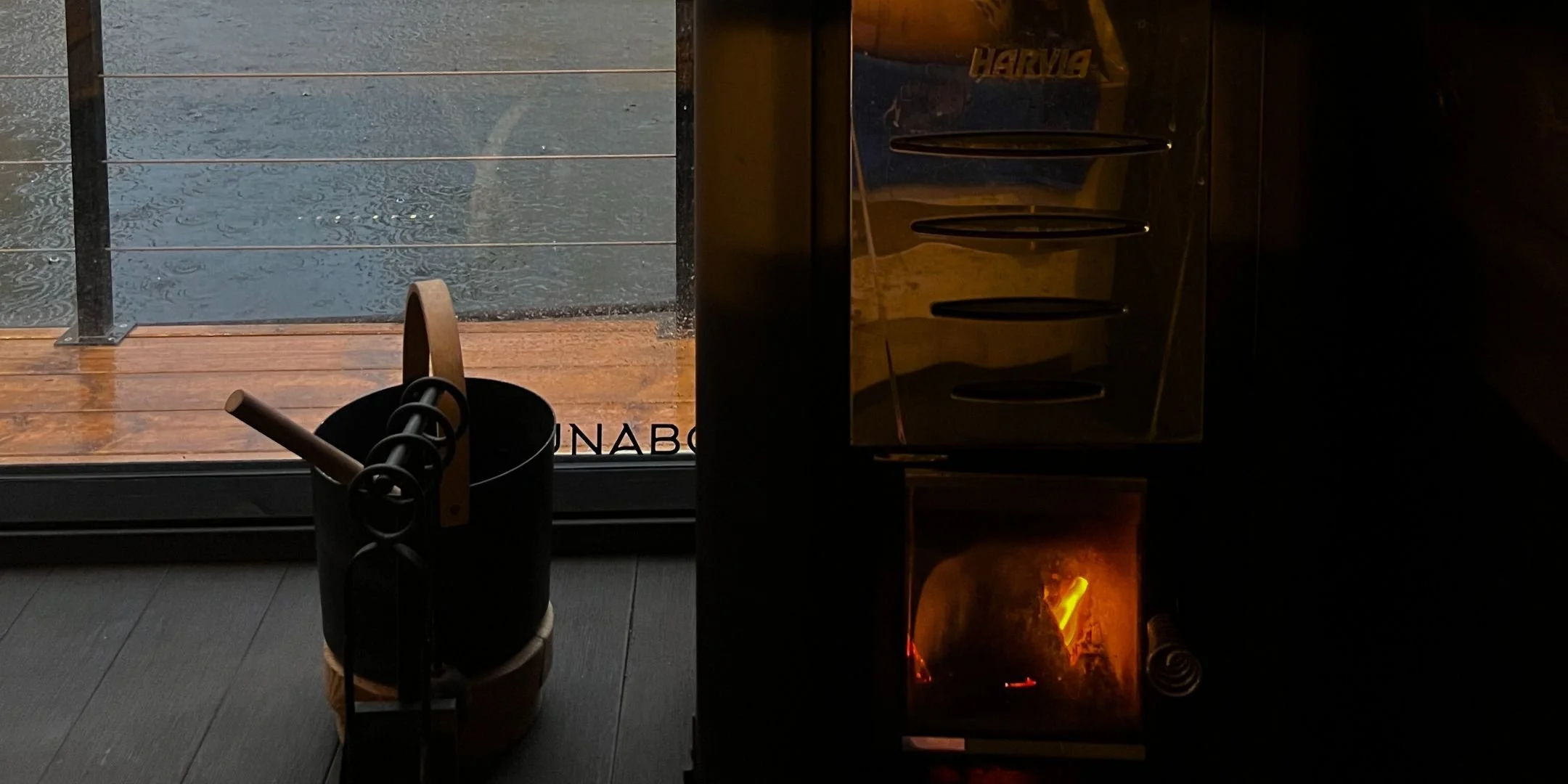a woodfired sauna stove and traditional bucket next to it, with a partial view of decking and water in the rain.