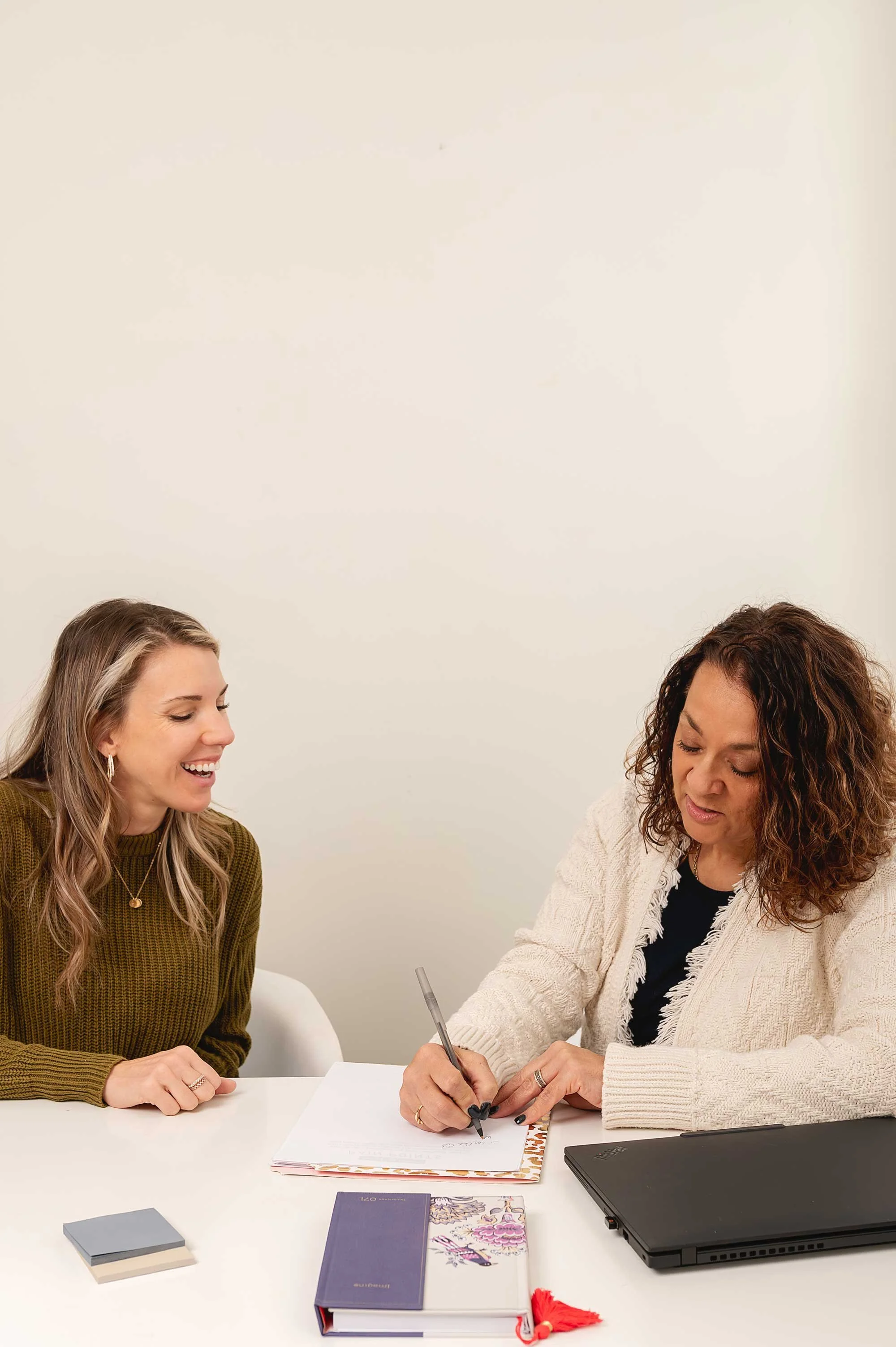 Two women sitting at a white table, one writing in a notebook, smiling and talking, with a laptop, notebooks, and a deck of cards on the table, against a plain white wall.