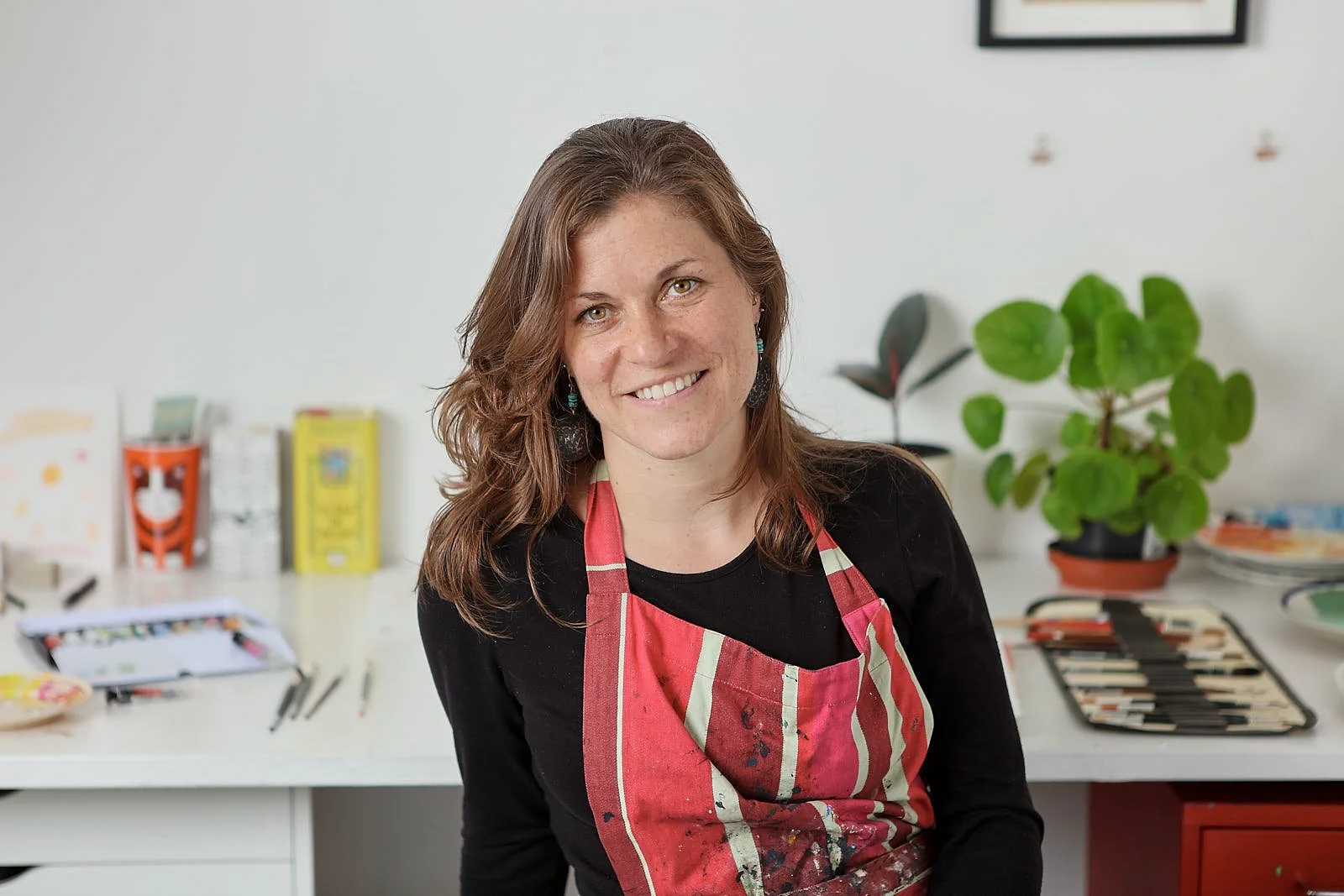 A photograph of Emma Dibben. She has shoulder length brown hair and is smiling at the camera. She is wearing a black top and a red and white striped apron with paint marks on it. The photograph is taken in her studio.