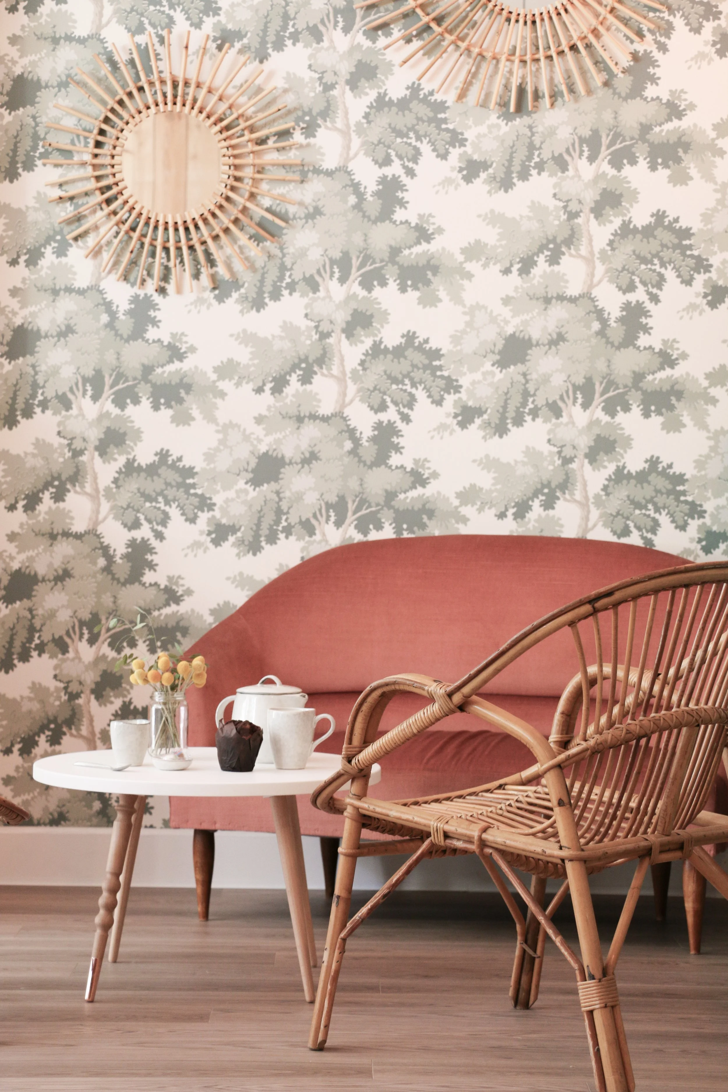 Un intérieur décoratif avec un canapé rose saumon, une table basse blanche avec des tasses, une théière, une petite bouteille avec des fleurs, une cupcake, un fauteuil en rotin, un mur à motif d'arbres en arrière-plan, et deux décorations rondes en bois pendant suspendues au plafond.