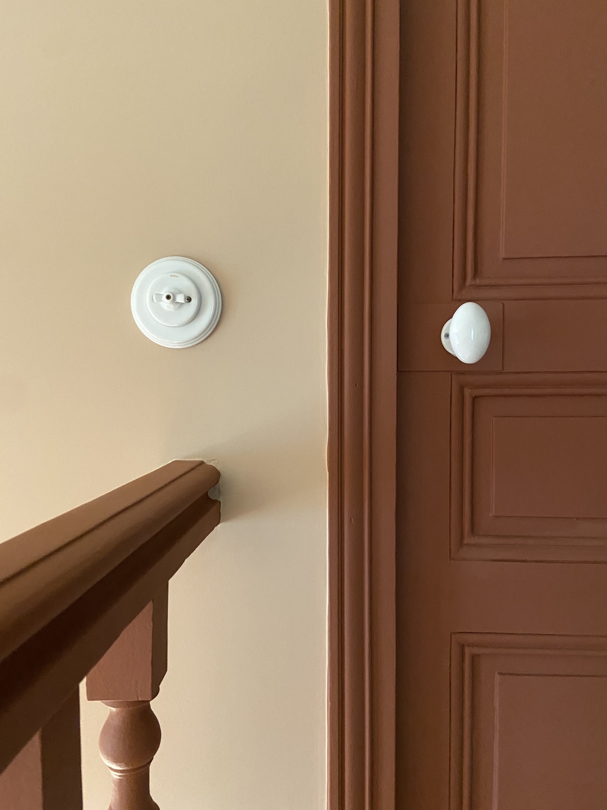 Détail d'une porte en bois moulurée et d'un escalier en bois peints en rouge calciné sur un mur rose doux et d'un interrupteur et d'une poignée de porte en porcelaine blanc