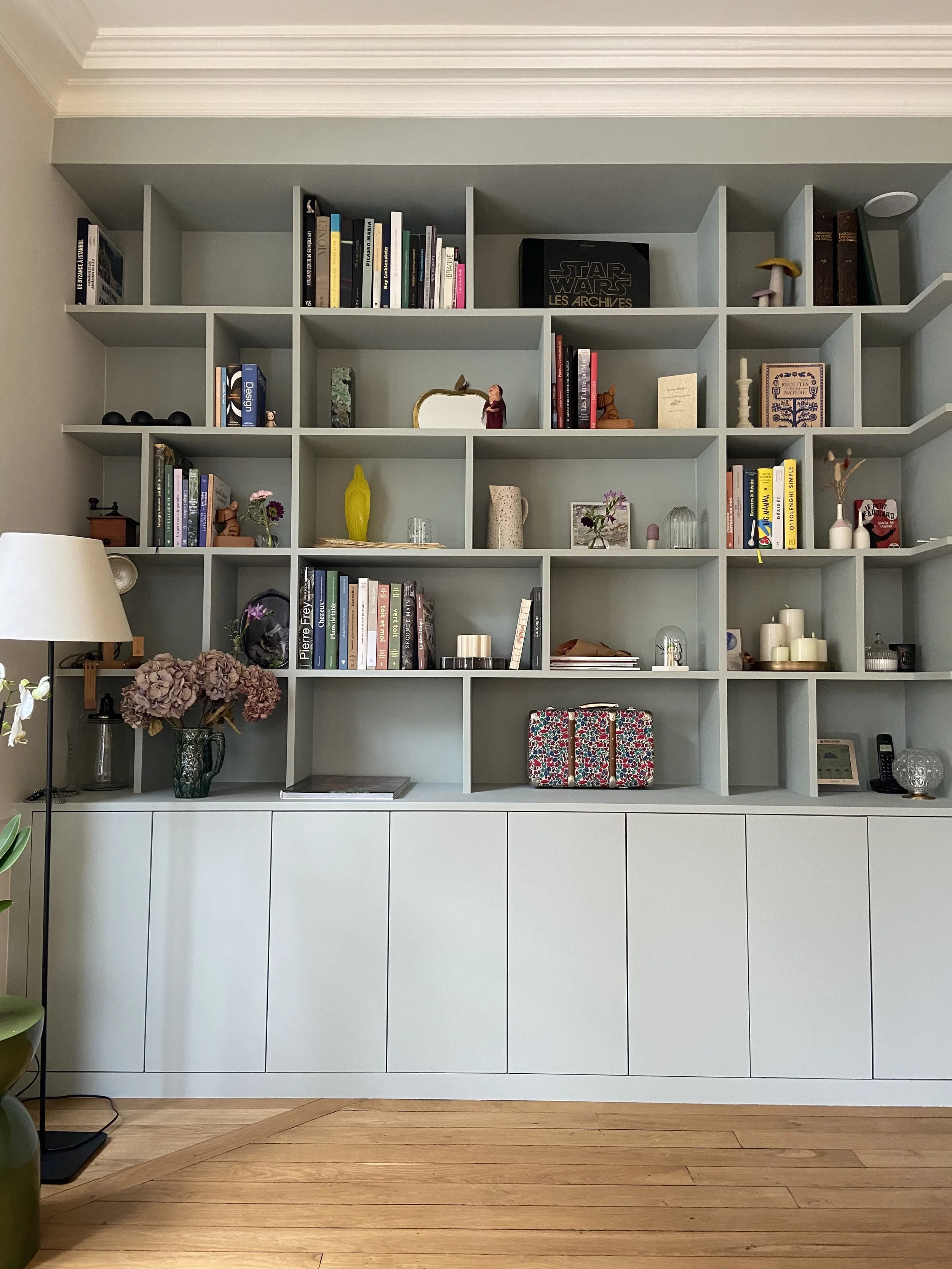 Grande bibliothèque murale remplie de livres, objets décoratifs et accessoires dans un salon, avec un placard bleu gris en dessous et un sol en parquet ancien