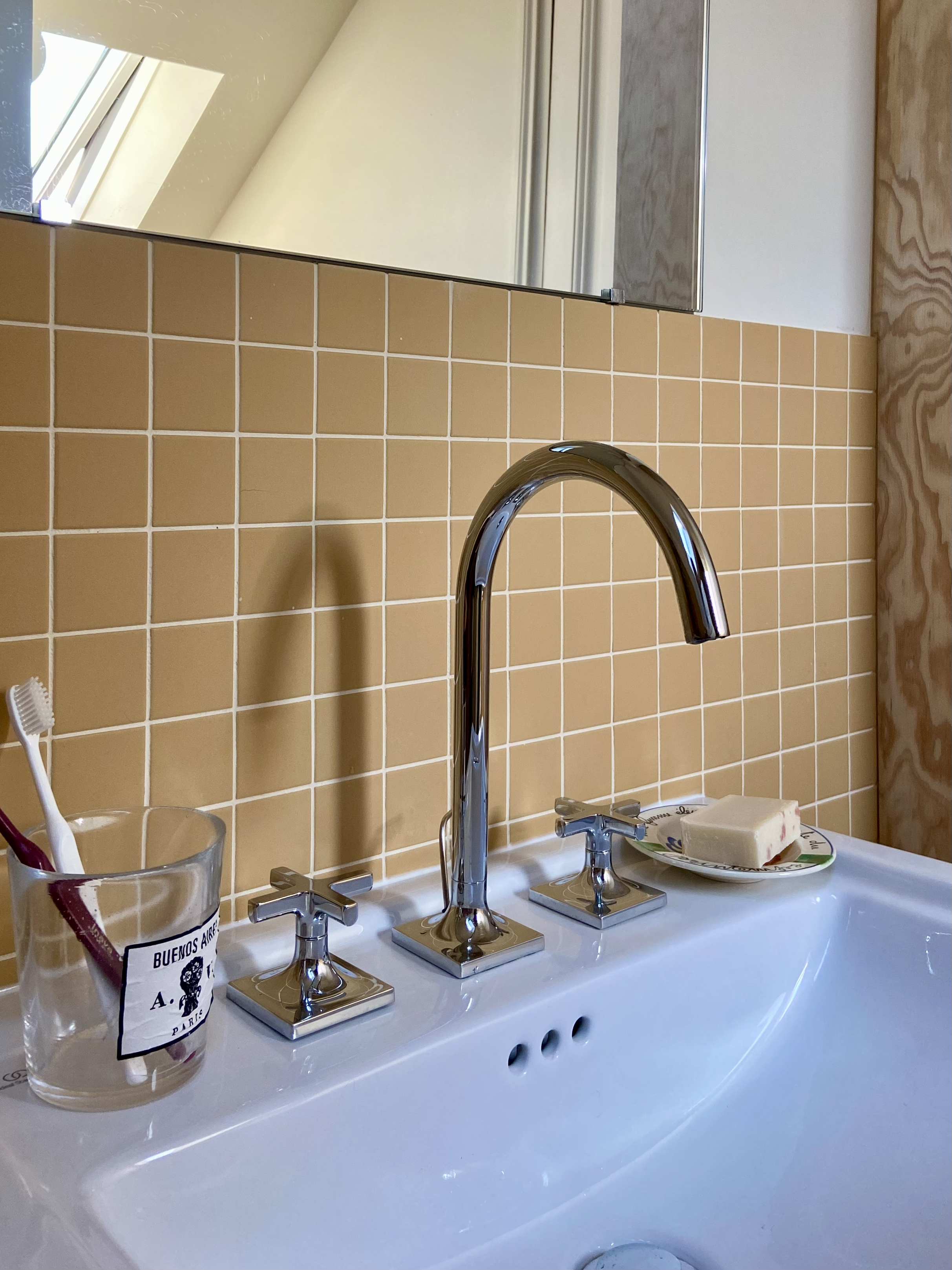 Détail d'un lavabo en céramique blanc vintage avec robinet en inox coudé et mélangeurs en croix, murs carrelé de petits carreaux jaunes et porte savon chiné