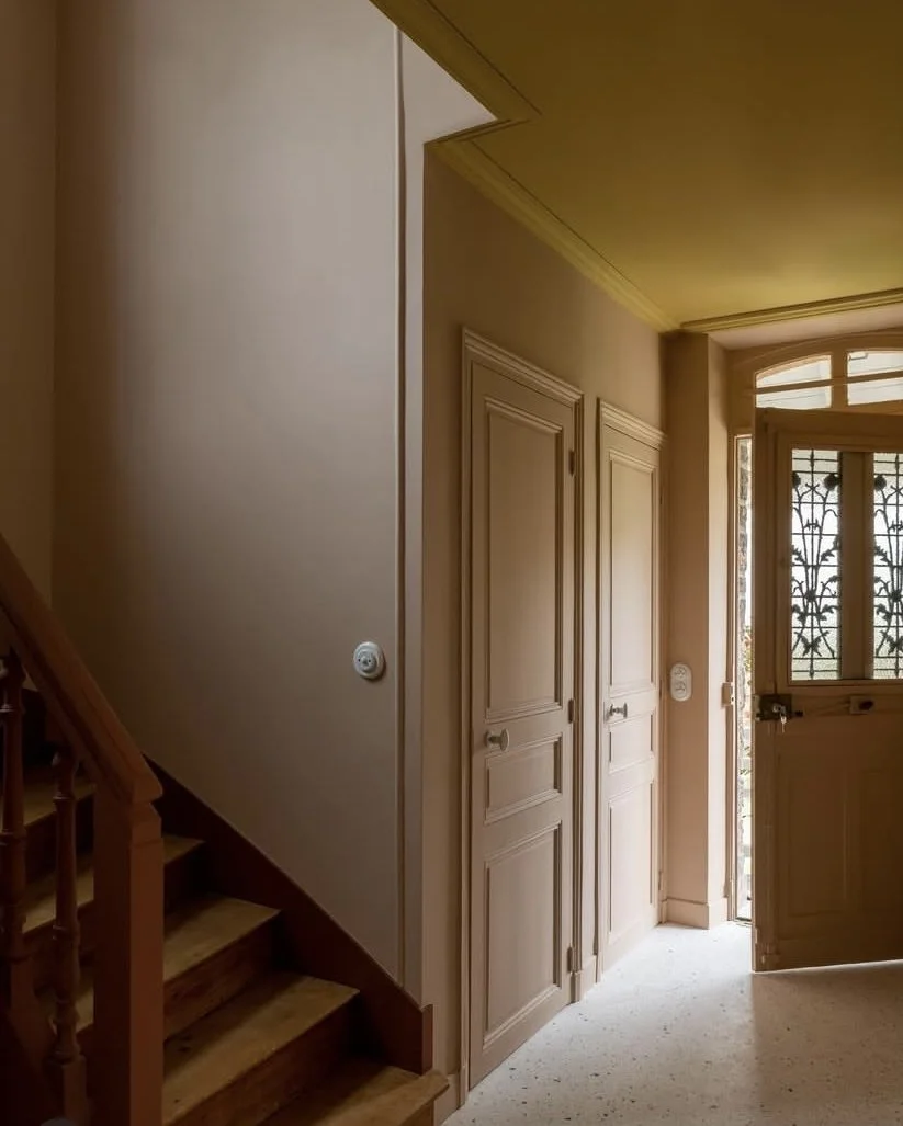 Entrée d'une maison meulière 1900 dans des teintes de rose, jaune indien et rouge calciné, moulures, terrazzo, escalier en bois massif, porte d'entrée avec fenêtre ouvragée