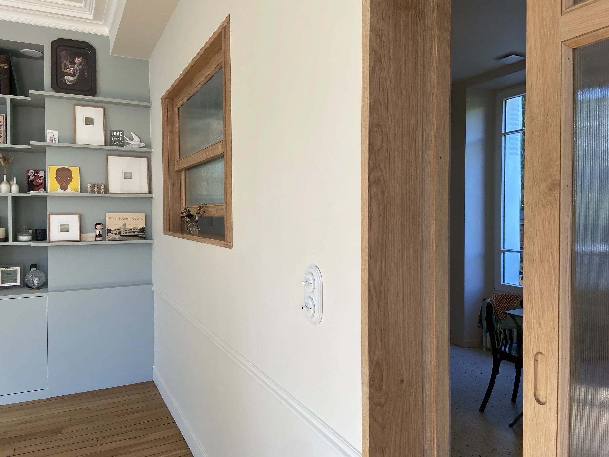 Détail d'un salon avec bibliothèque intégrée peinte en bleu gris "light blue" avec de multiples objets décoratifs, fenêtre en bois à guillotine et verre fluté, interrupteurs en porcelaine, moulures