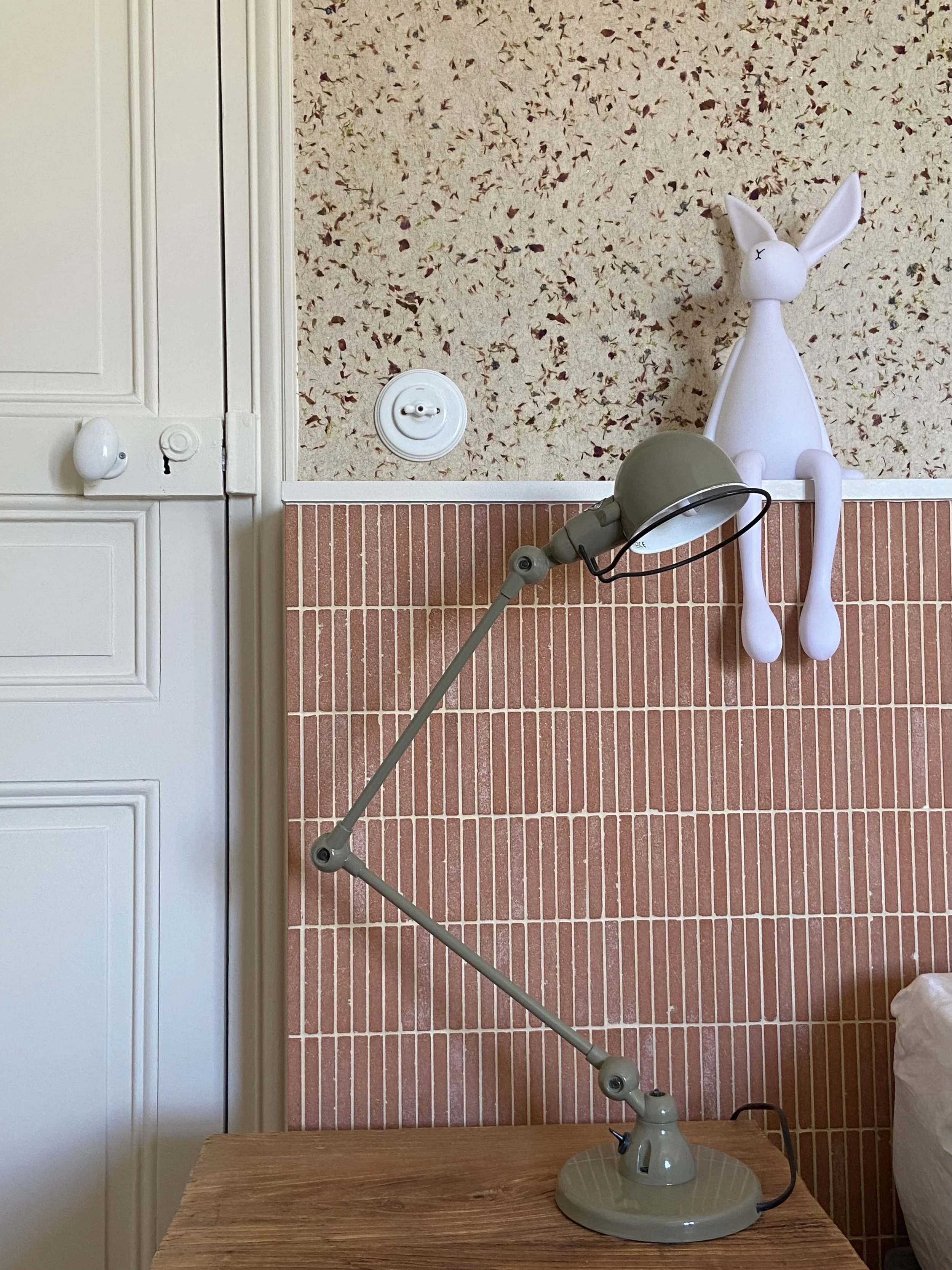 Détail d'une tête de lit carrelée en briquette, papier peint de fleurs séchées, lampe Jieldé, lampe lapin et interrupteur en porcelaine