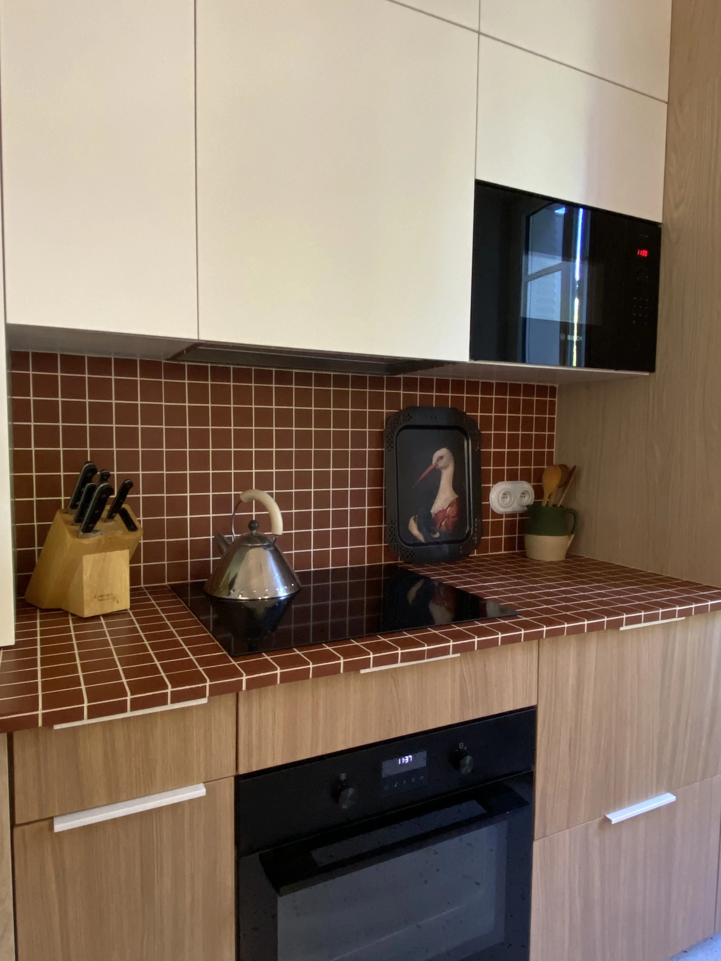 Cuisine façades bois et blanches avec plaques de cuisson, four, micro-ondes, crédence et plan de travail en petits carreaux bordeaux, couteaux dans un bloc, théière en métal, décoration avec un tableau d'oie, pot avec cuillères en bois.